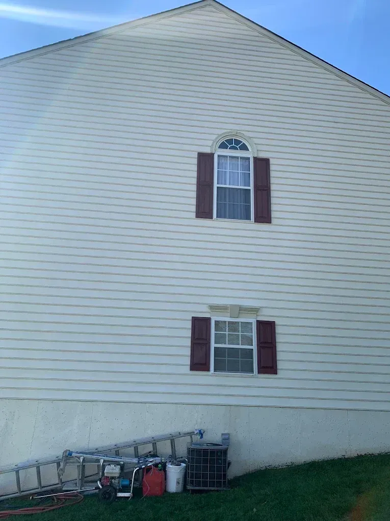 White house side wall with two small windows, maroon shutters, and items stored along the foundation.