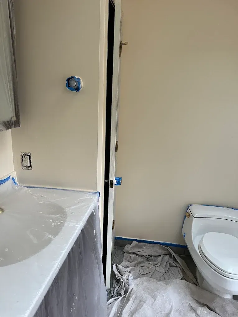Bathroom under renovation with exposed wall edge, sink, and toilet protected by plastic and tape.