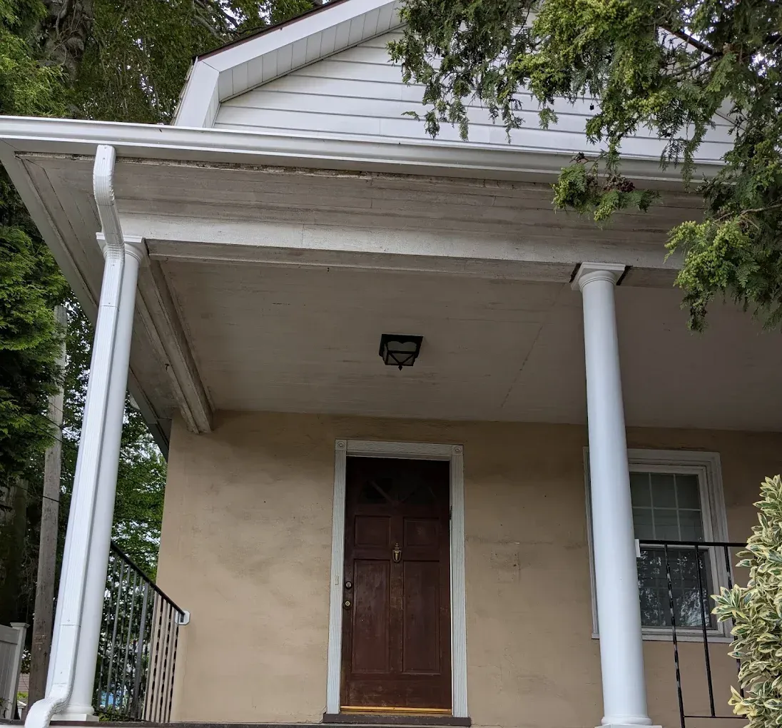 Front porch of a beige house with white columns and a brown door, viewed from below.