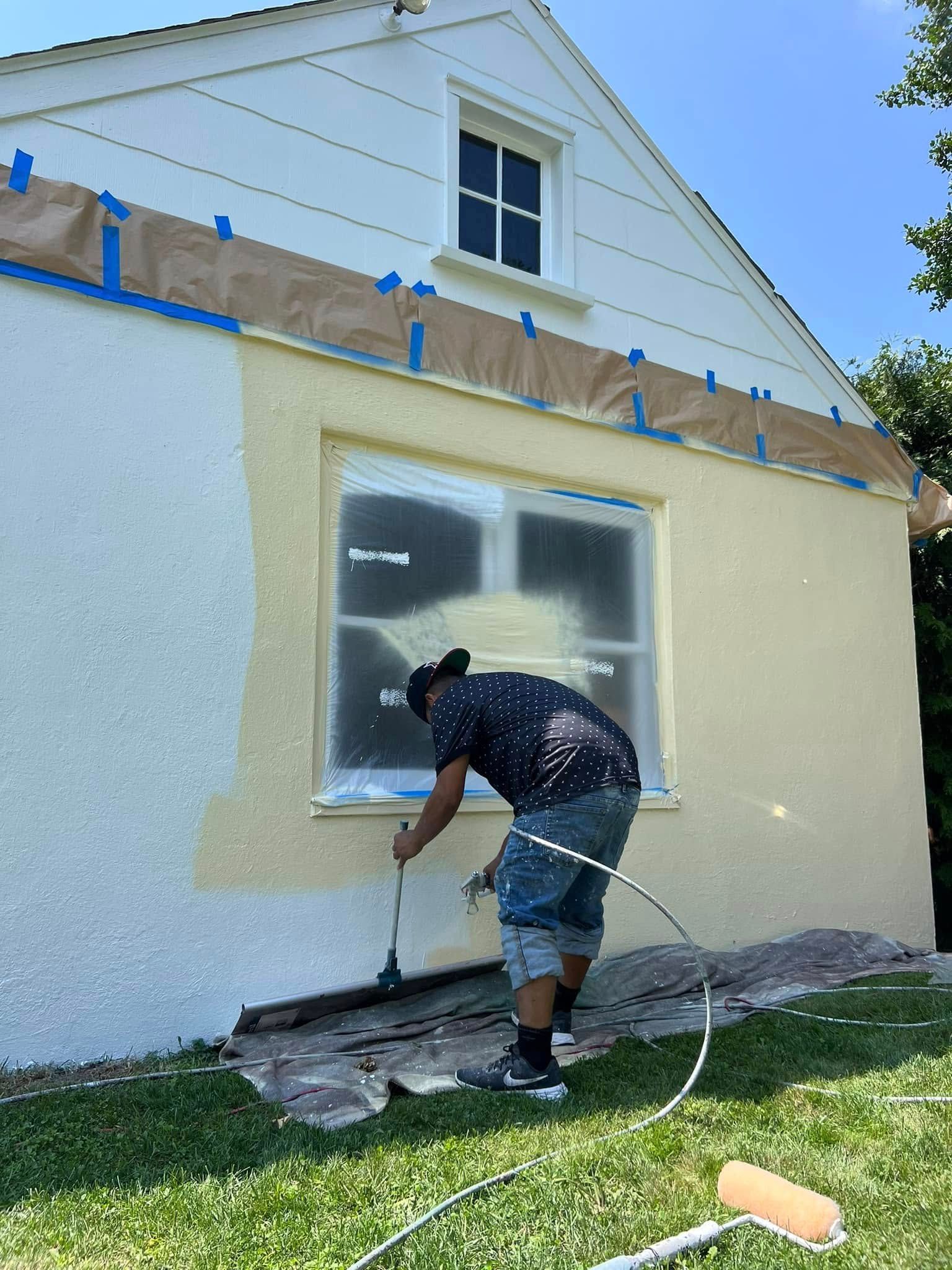 Worker spray-painting the lower exterior wall of a house, with windows and taped trim above.