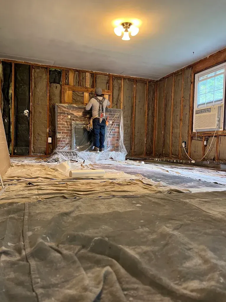 Renovation in progress in an empty room with exposed studs, plastic-covered floor, and a person at the wall unit
