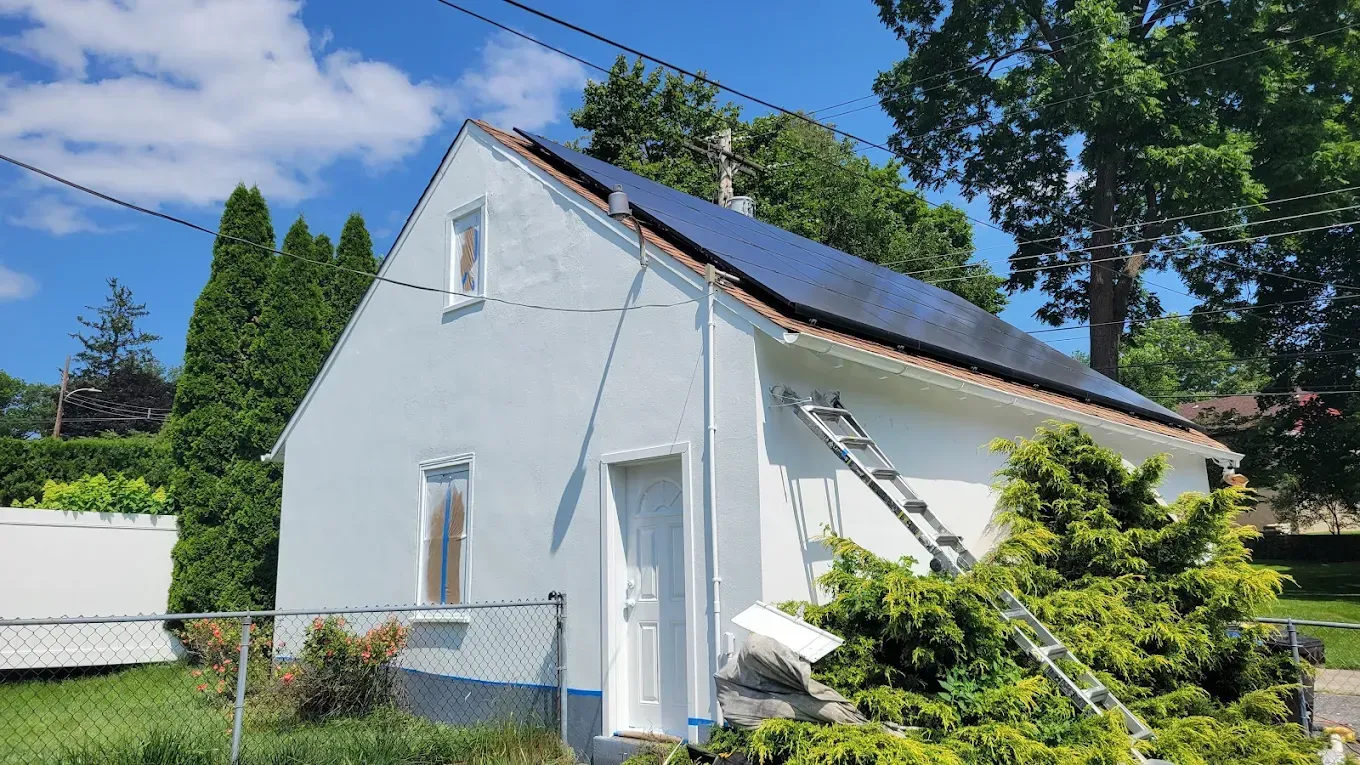 White house with steep roof and solar panels, surrounded by green trees under a bright blue sky