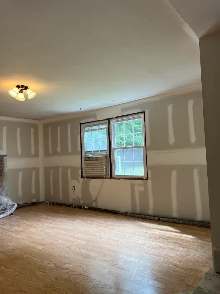 Empty beige room with unfinished drywall, wood floor, ceiling light, and a window AC unit by the window