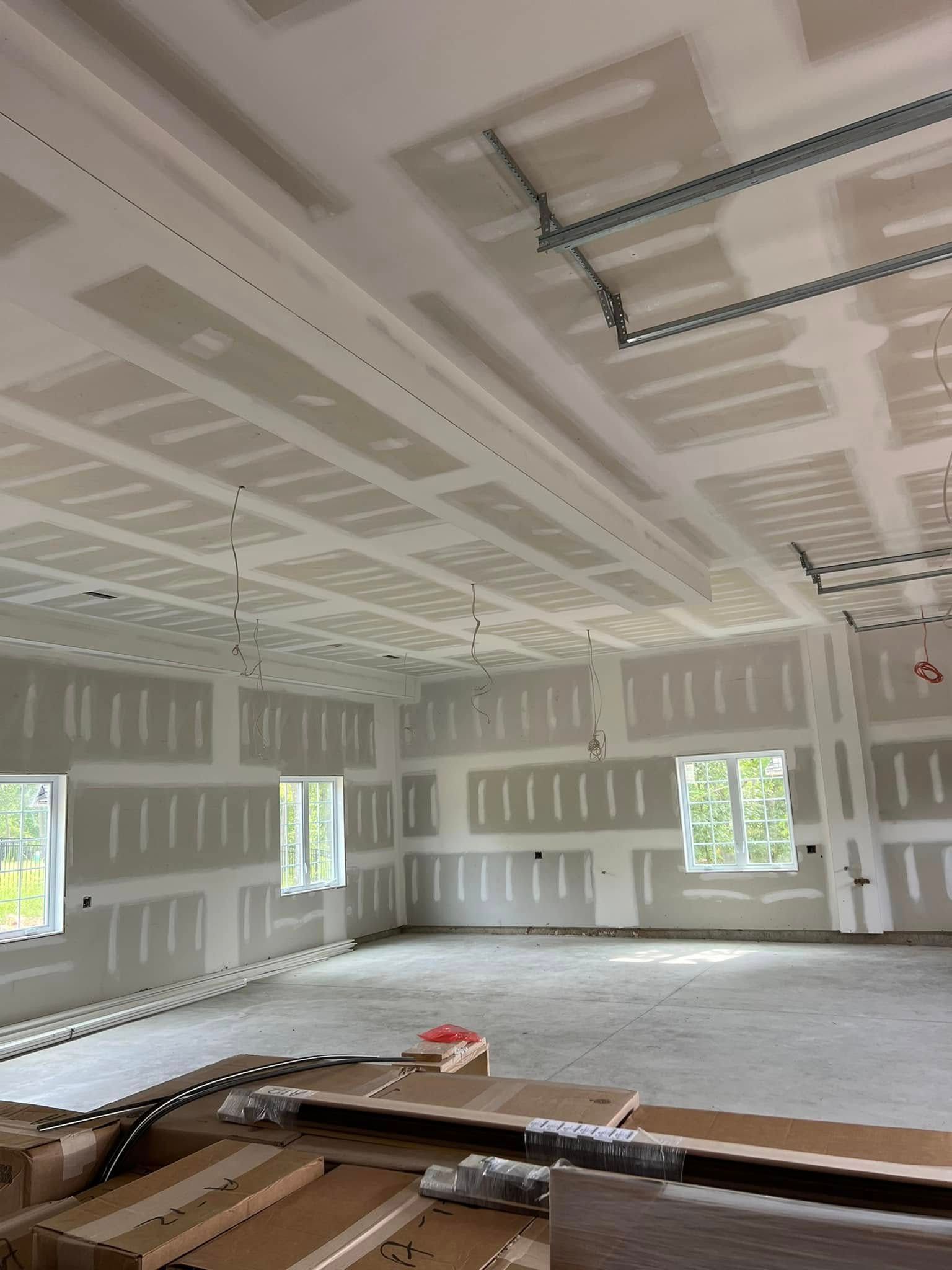 Empty drywall-lined room under construction with windows, ceiling joints, and ladders/materials on the floor