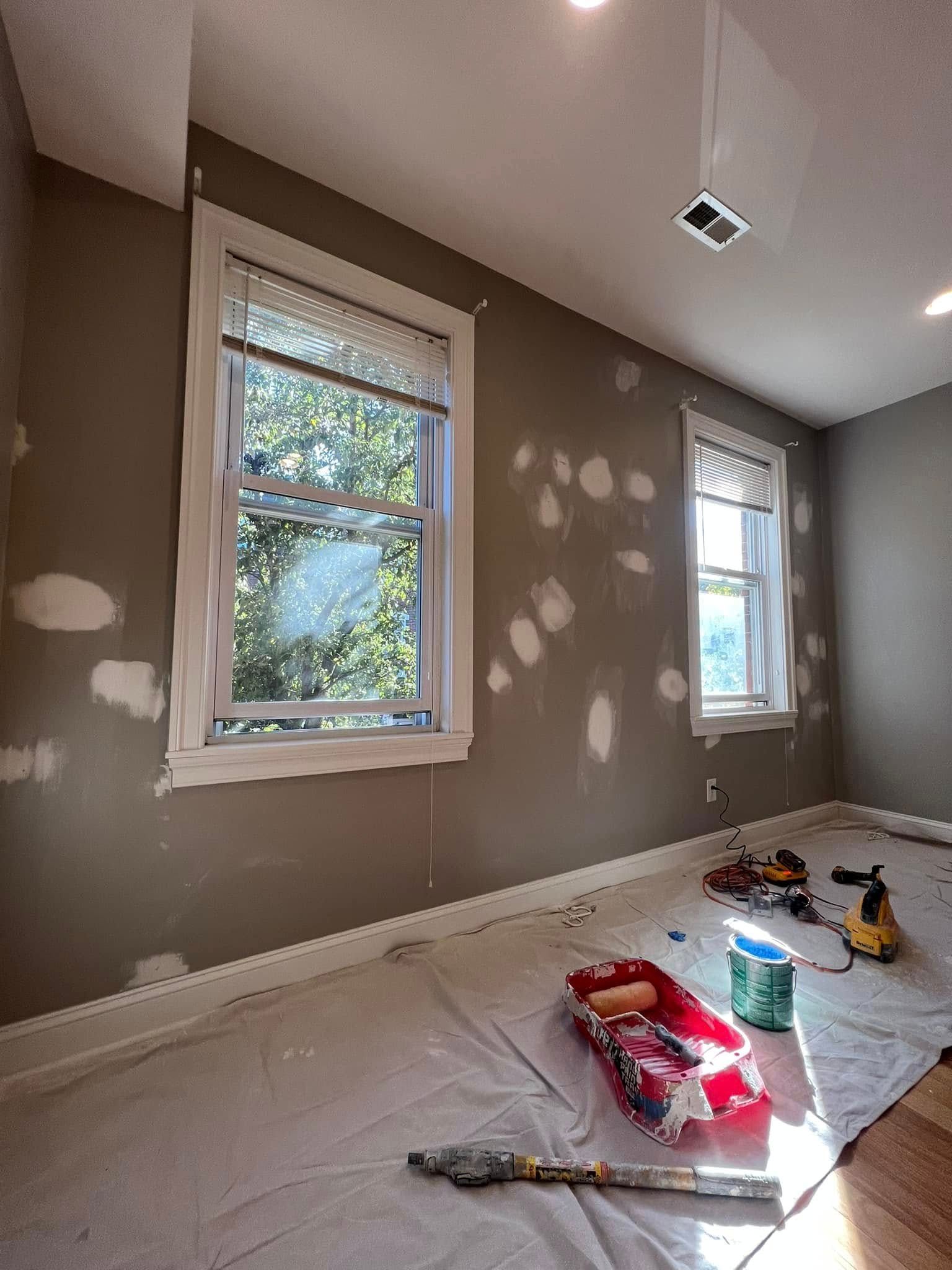 Painted room with two windows, patchy wall repair spots, and paint supplies on a drop cloth over a hardwood floor