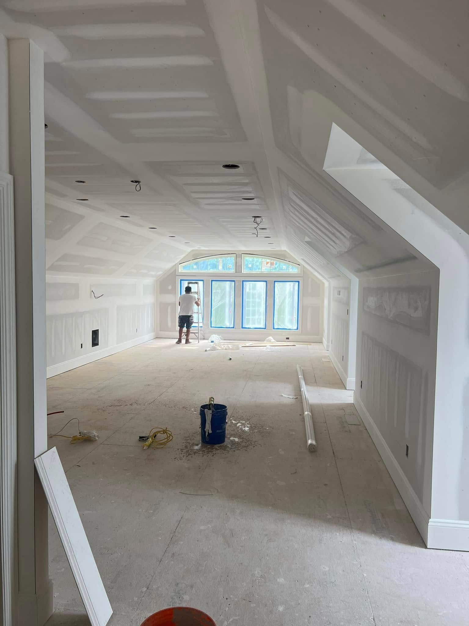 Unfinished white attic room with drywall, scattered debris, and a worker near the far windows.