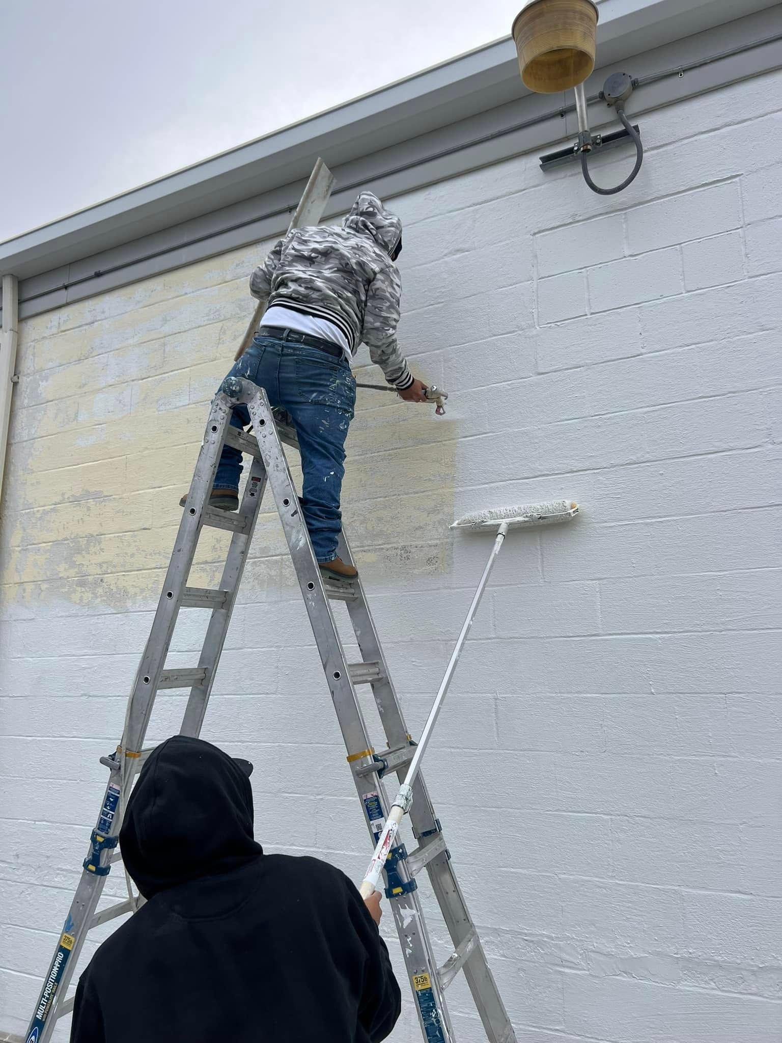 Person on a ladder painting an exterior white wall while another stands nearby.