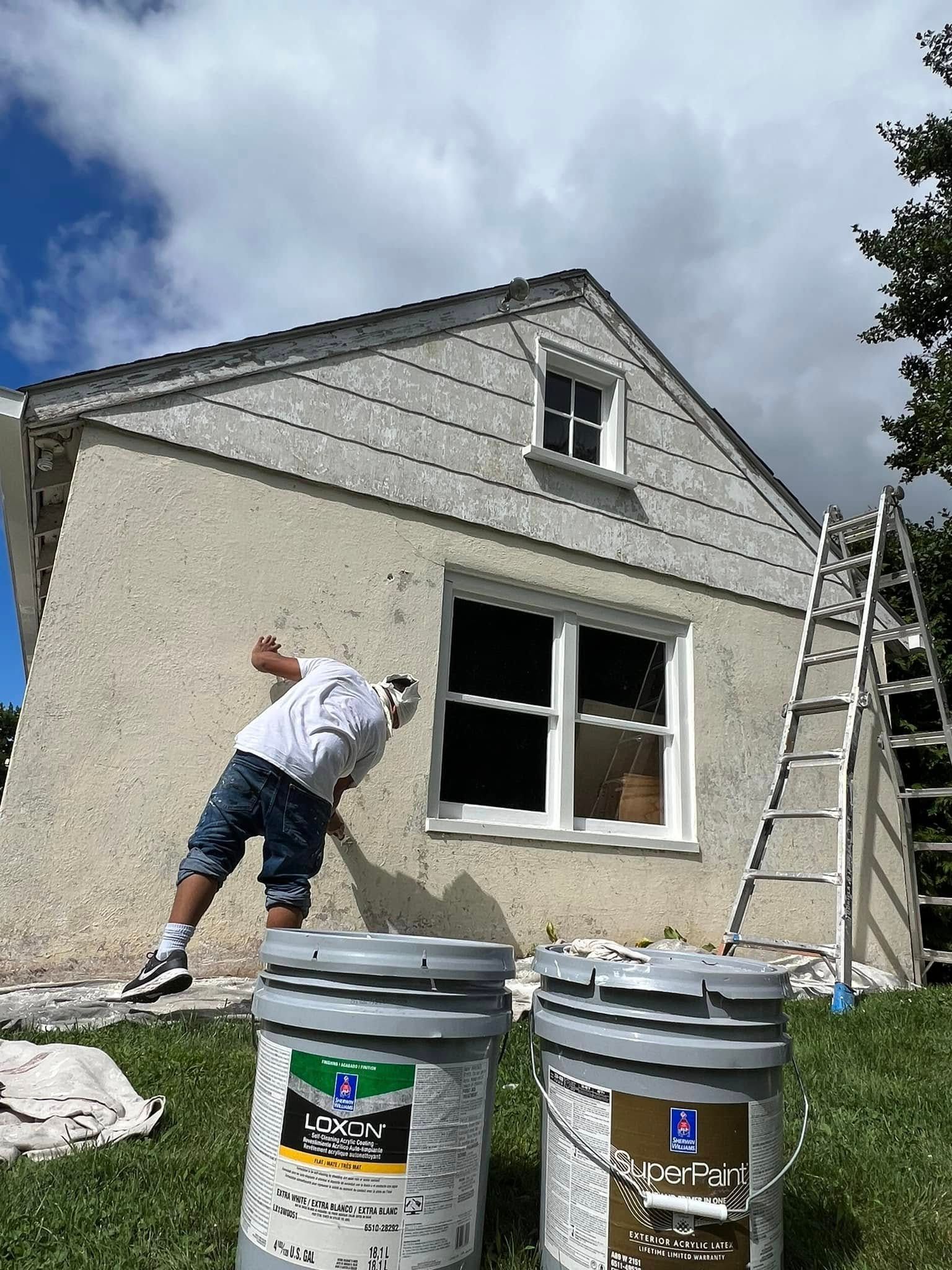 Worker painting a house exterior near a ladder and buckets under a cloudy sky