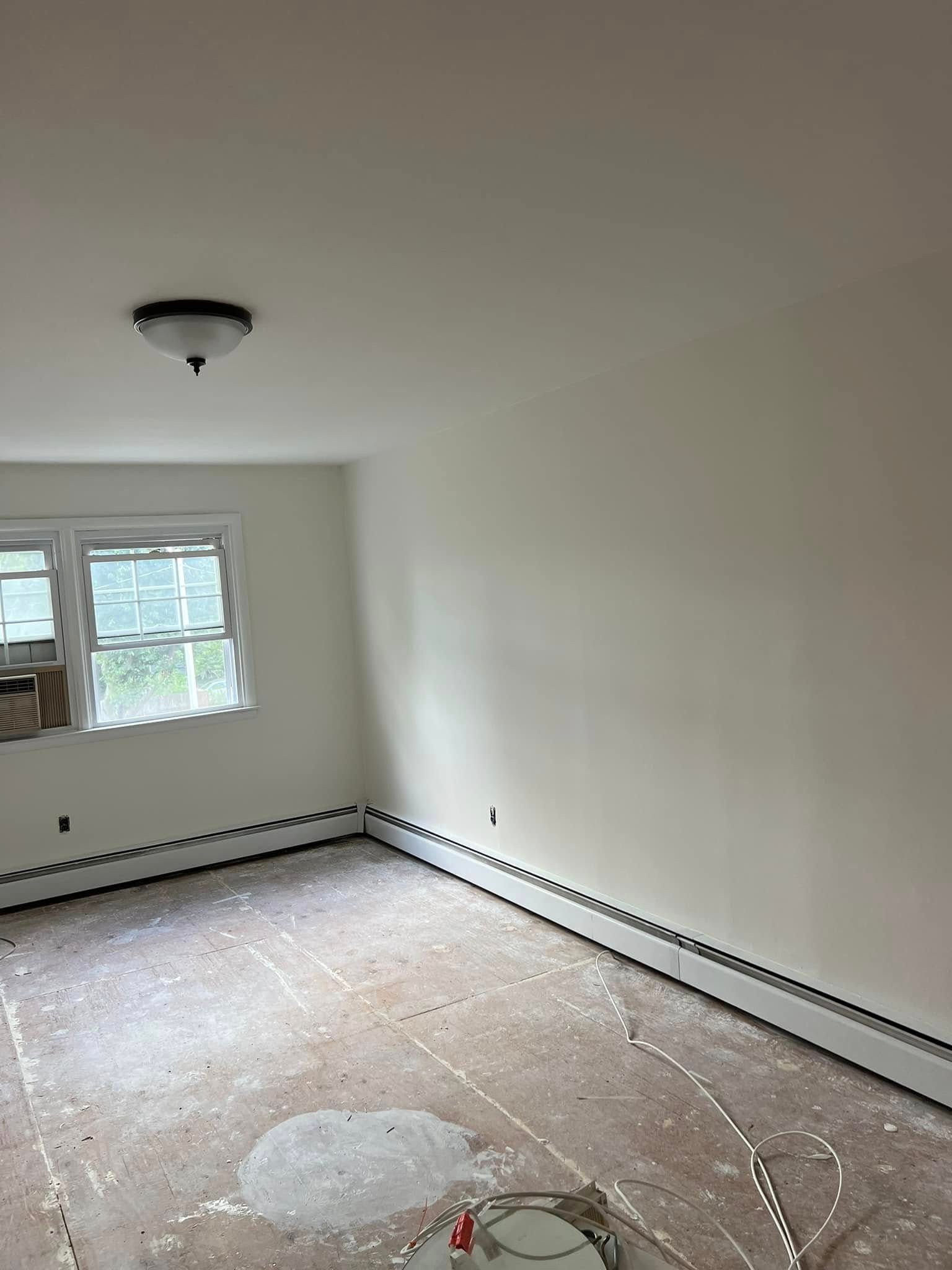 Empty room with white walls, a window, ceiling light, and unfinished floor under renovation