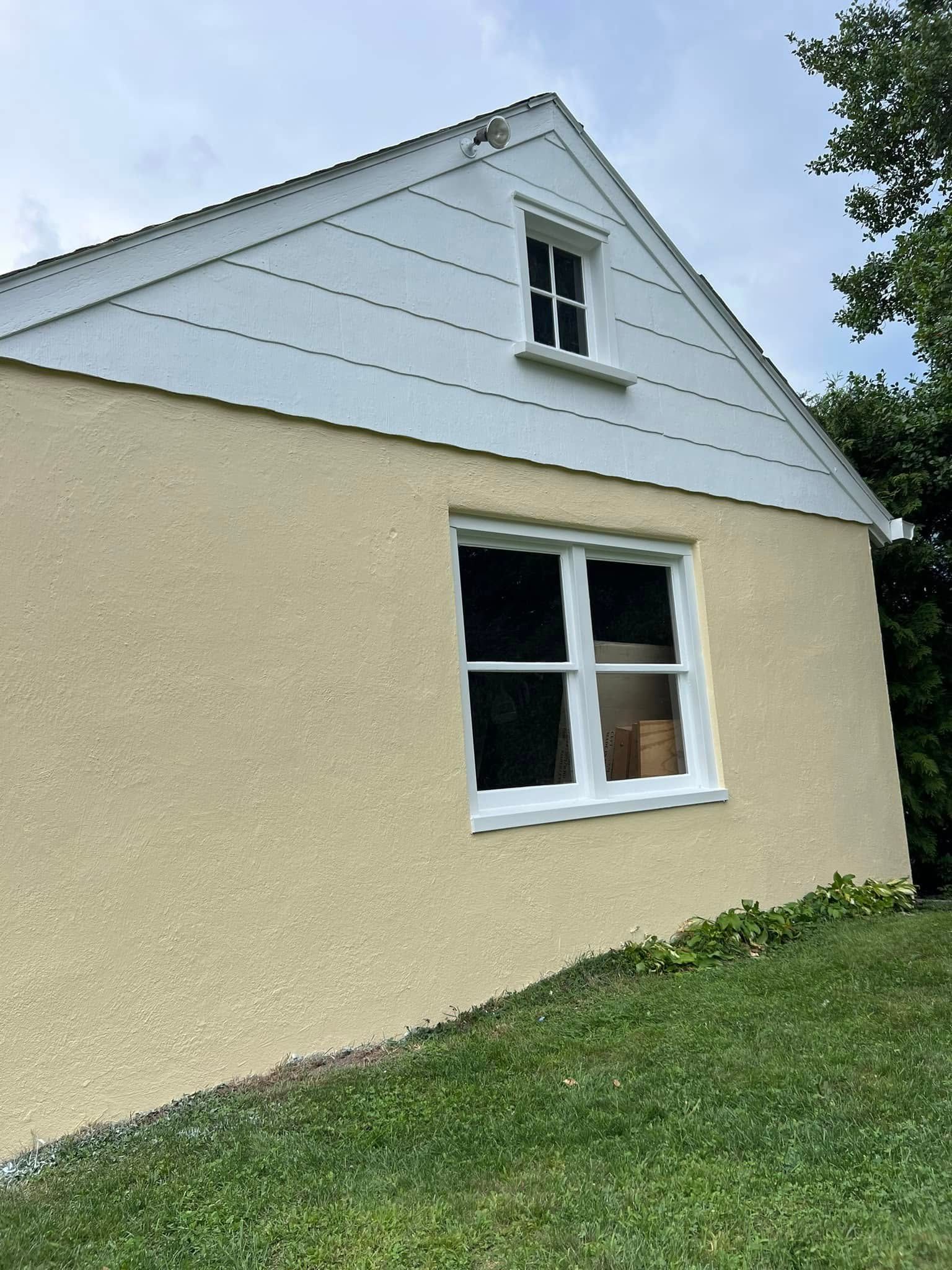 Small beige house with white siding gable and a white-framed window on a grassy slope