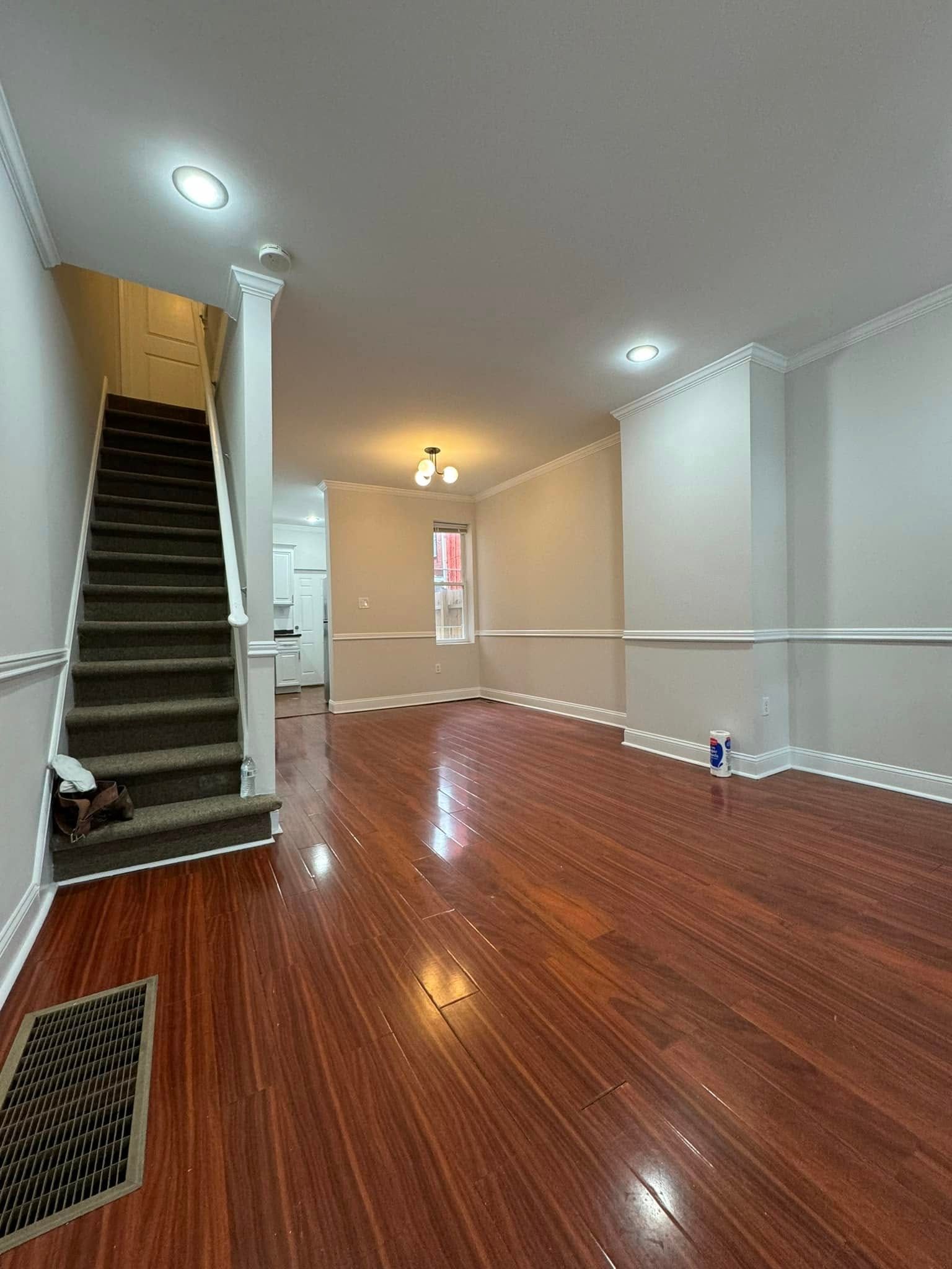 Empty hallway with glossy wood floors, beige walls, stairs on the left, and recessed ceiling lights