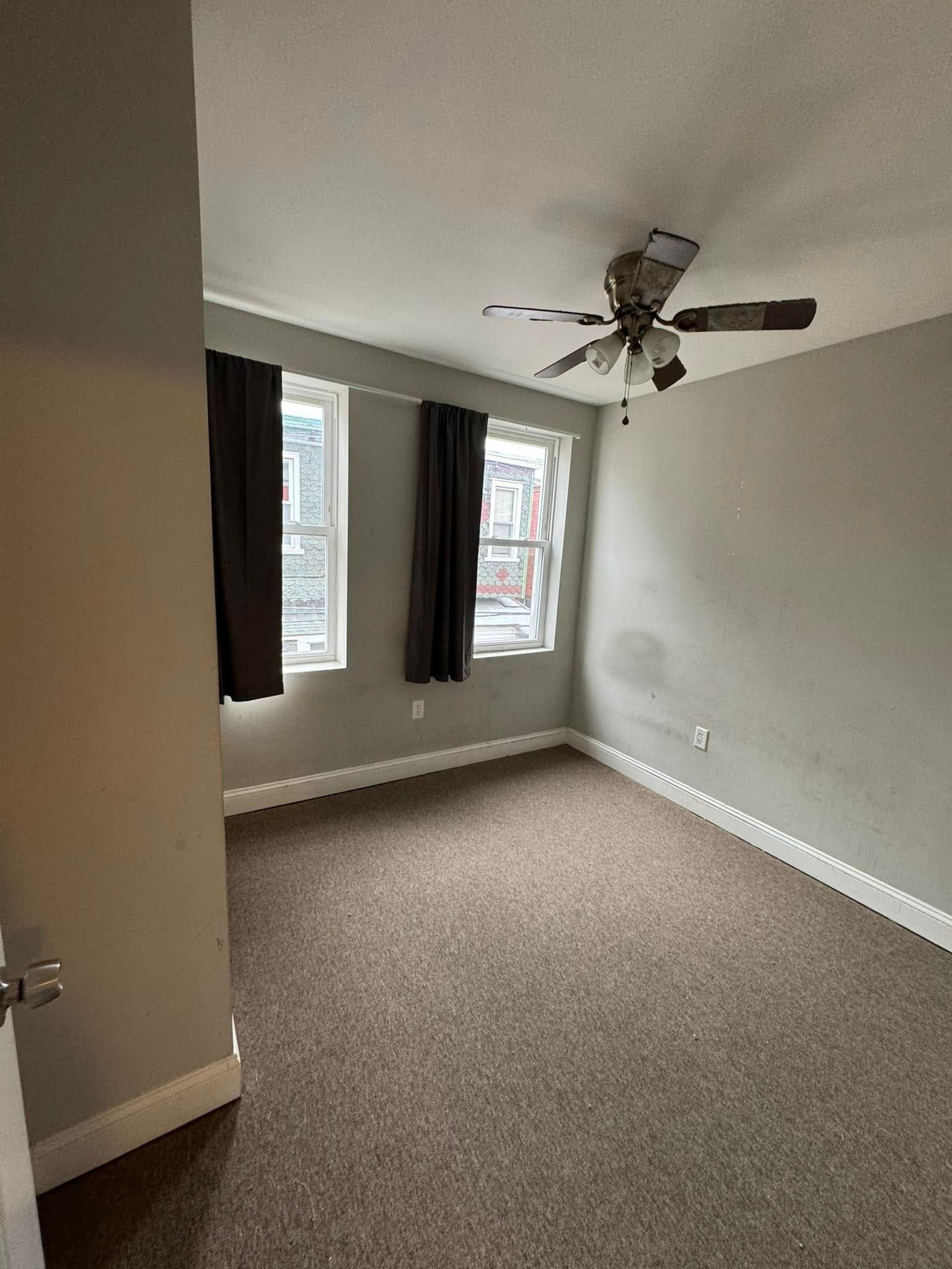 Empty beige bedroom with carpet, two windows with curtains, and a ceiling fan