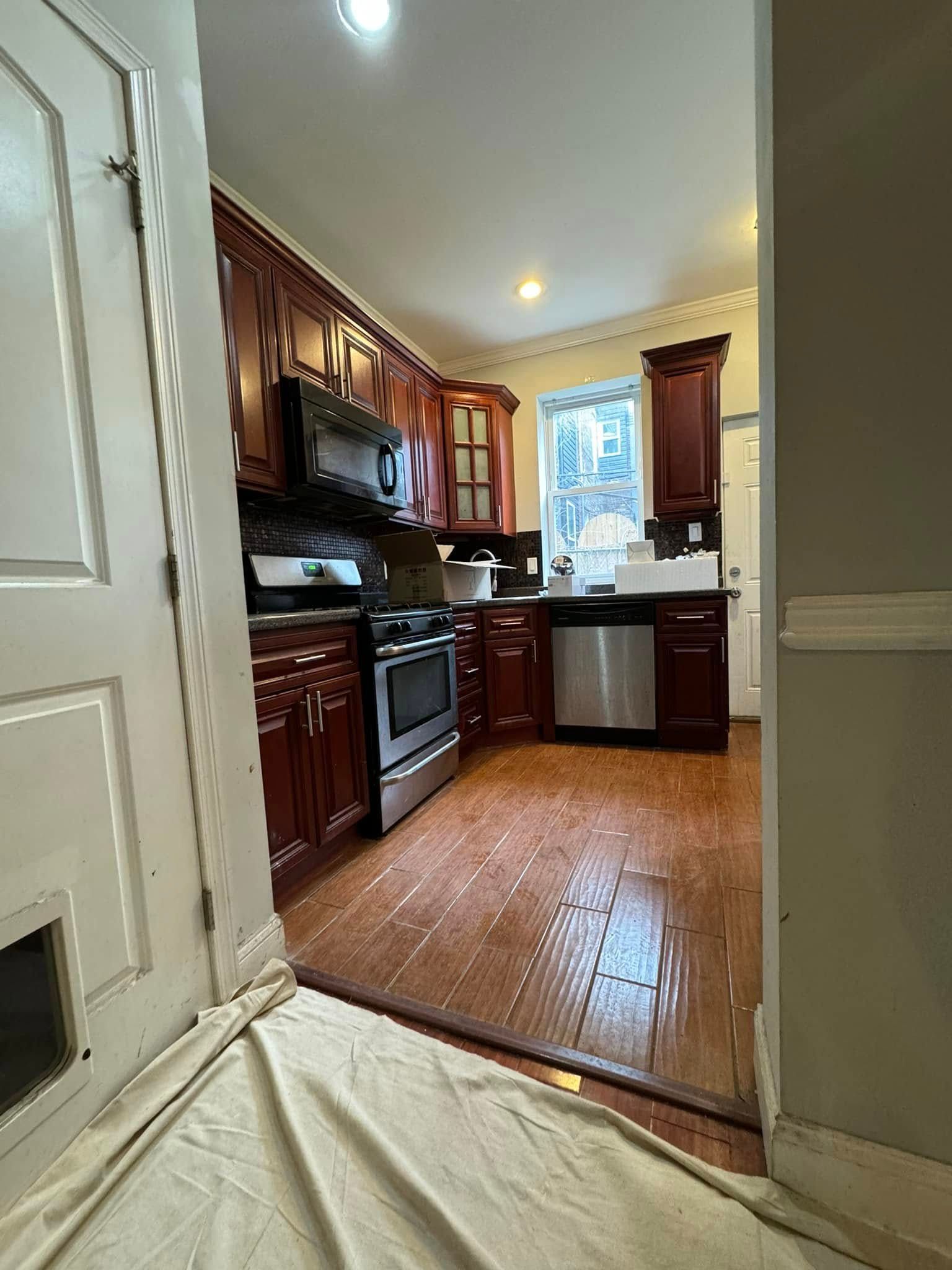 Narrow galley kitchen with dark wood cabinets, stainless steel appliances, and a window at the end.