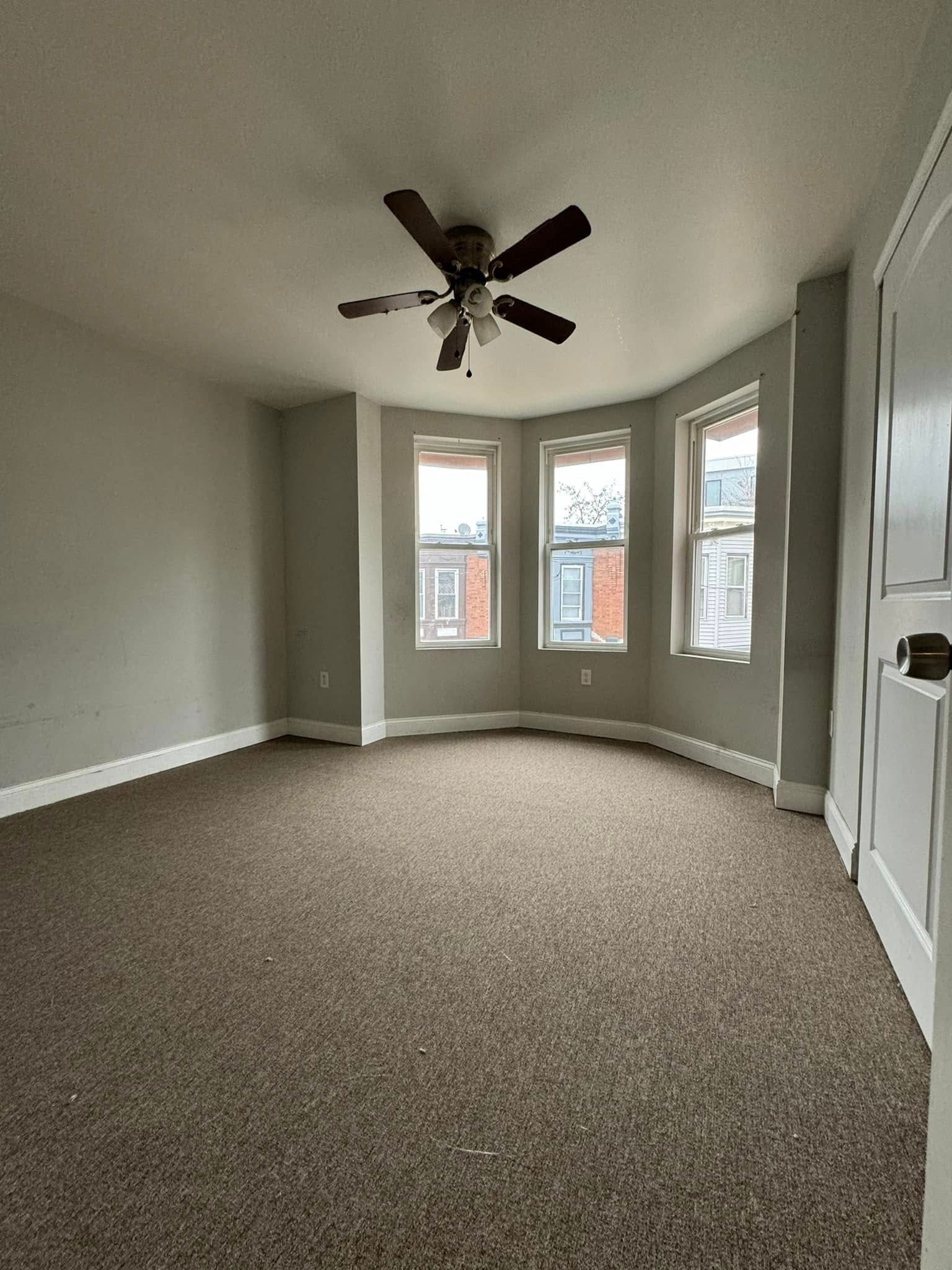 Empty carpeted room with bay windows, gray walls, and a ceiling fan.