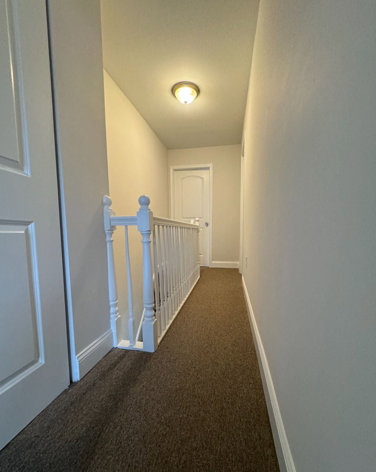 Narrow upstairs hallway with beige carpet, white walls, railing, and a door at the end.
