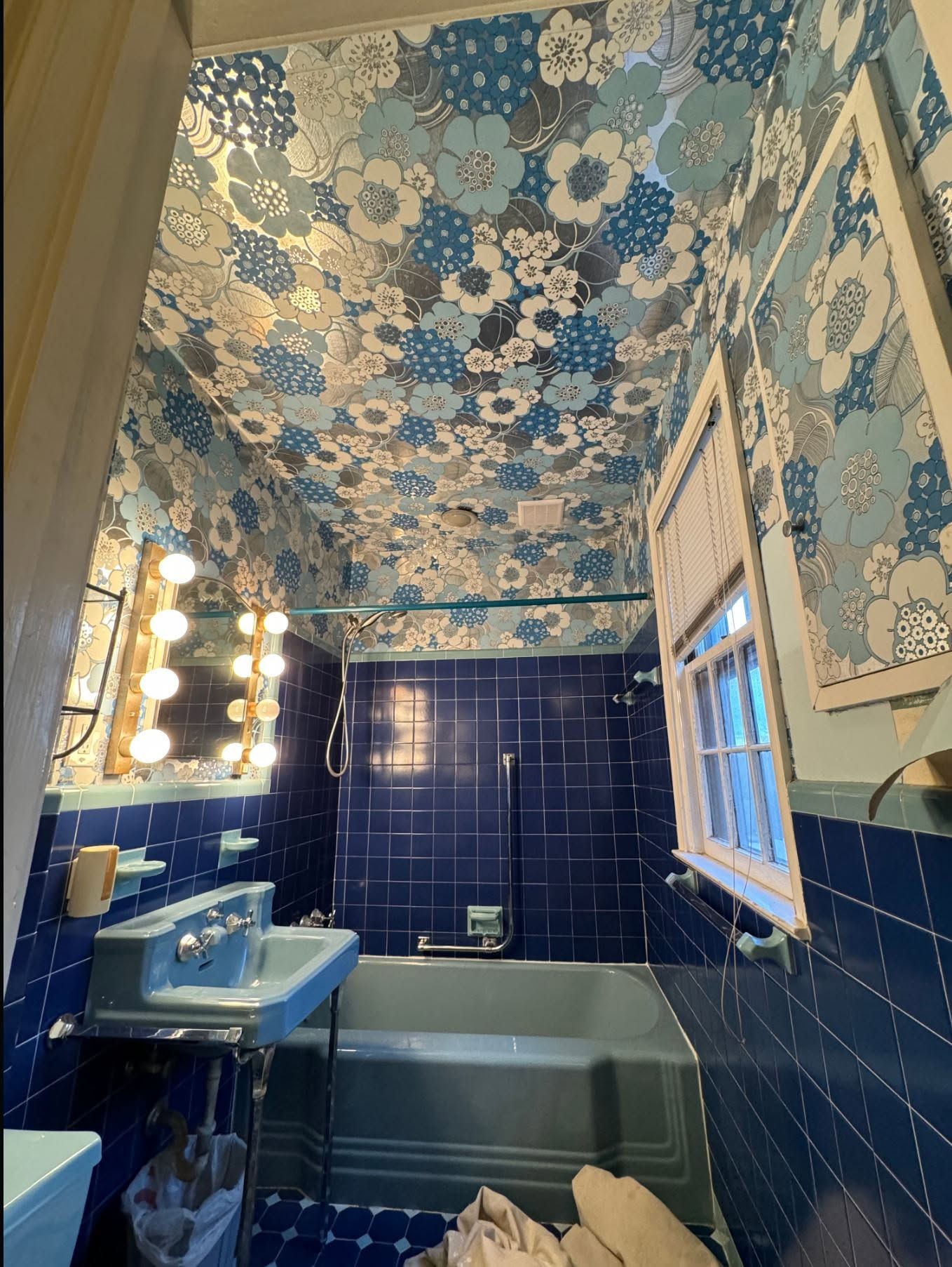 Blue-tiled bathroom with floral wallpaper, a sink, mirror lights, and a bathtub under a window.