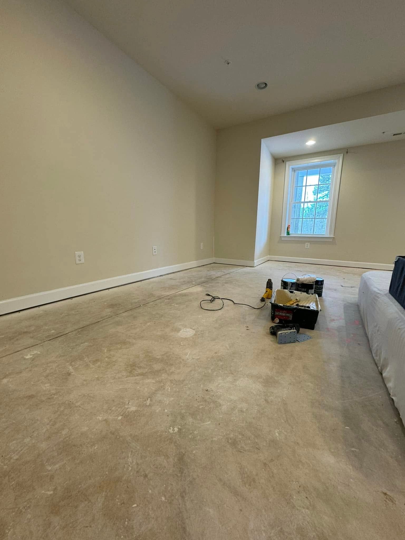 Empty beige room with bare carpet, window light, and cleaning supplies on the floor