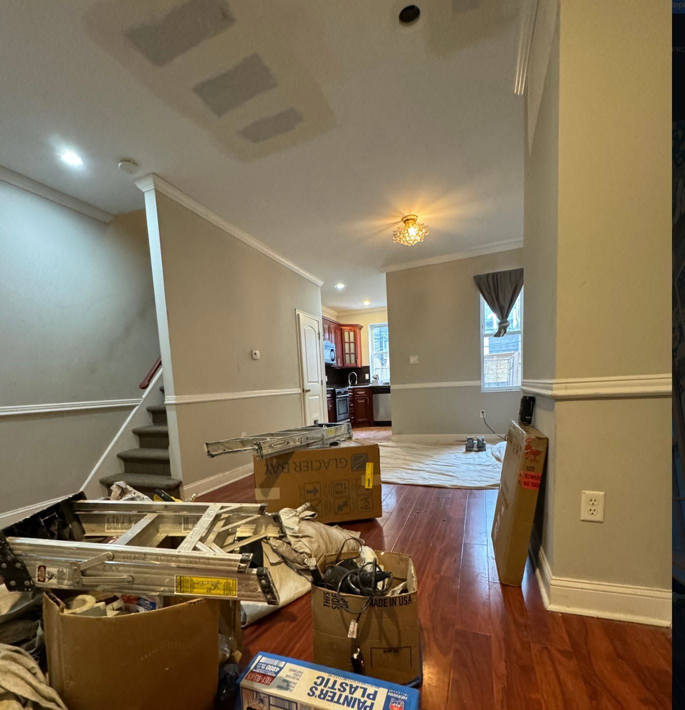 Cluttered living room with boxes, unpacked items, and furniture amid a hallway and hardwood floors.