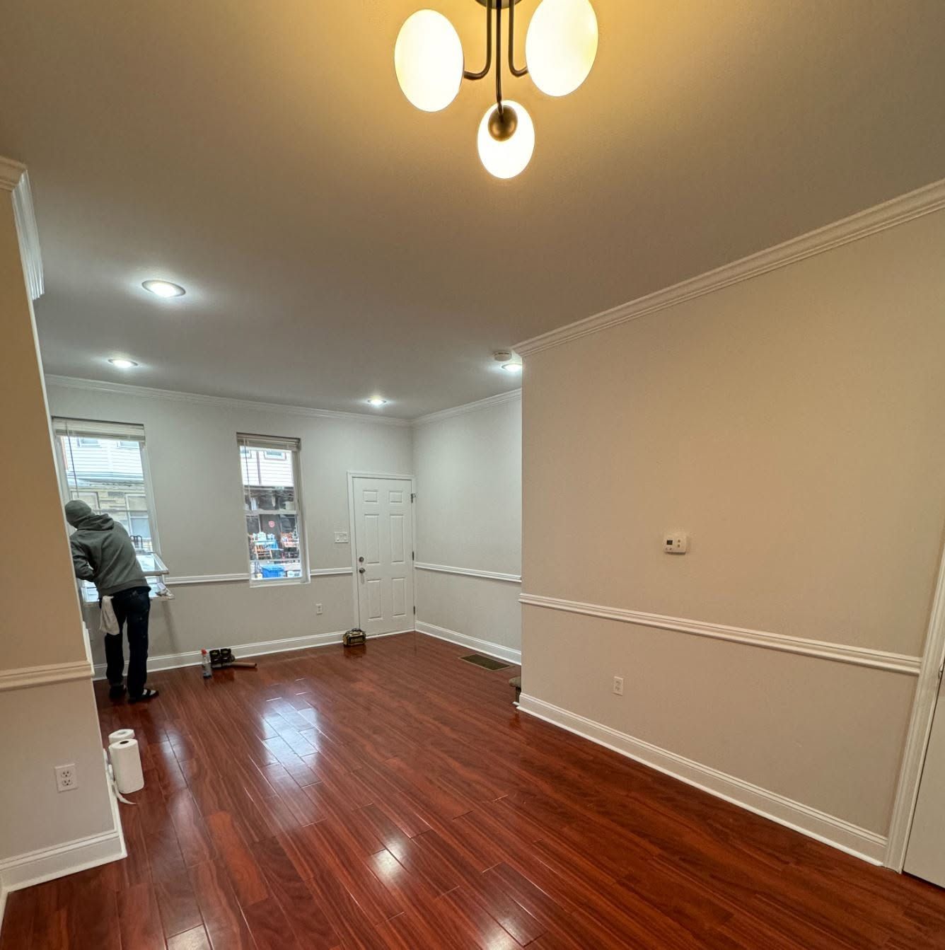 Empty beige room with glossy wood floor, ceiling lights, and a person near the far windows.