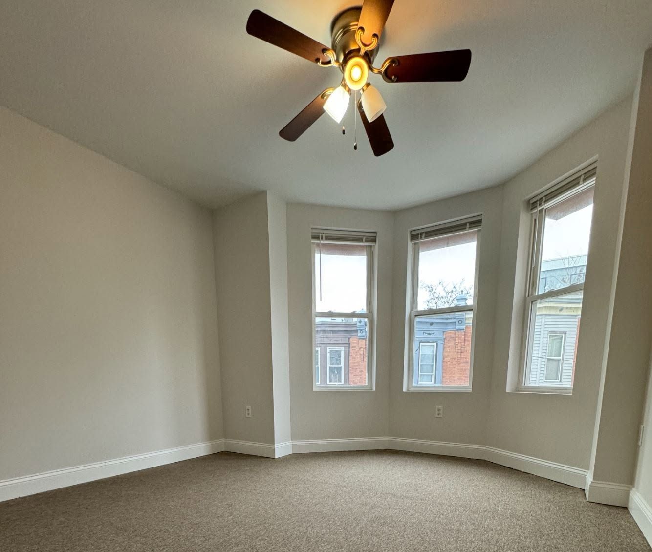 Empty carpeted room with bay windows, beige walls, and a ceiling fan lit overhead.