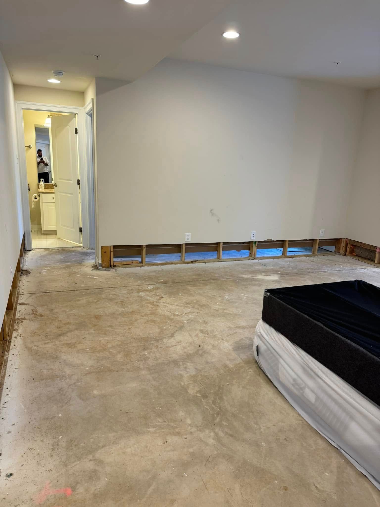 Unfinished bedroom with exposed floor, wall cutouts, and a bed at right; hallway visible in back.