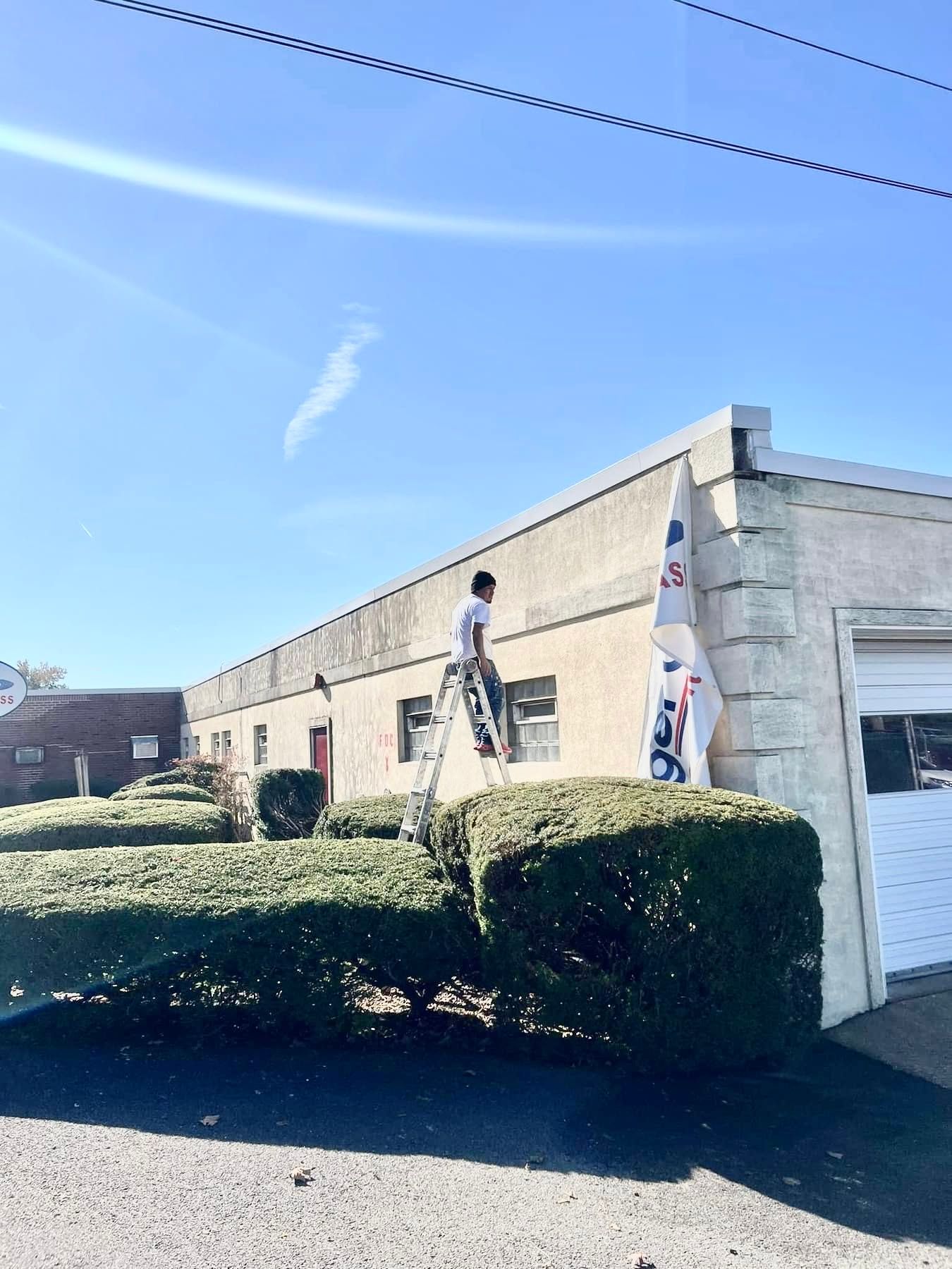 Person on a ladder cleaning a beige building’s exterior under a clear blue sky.