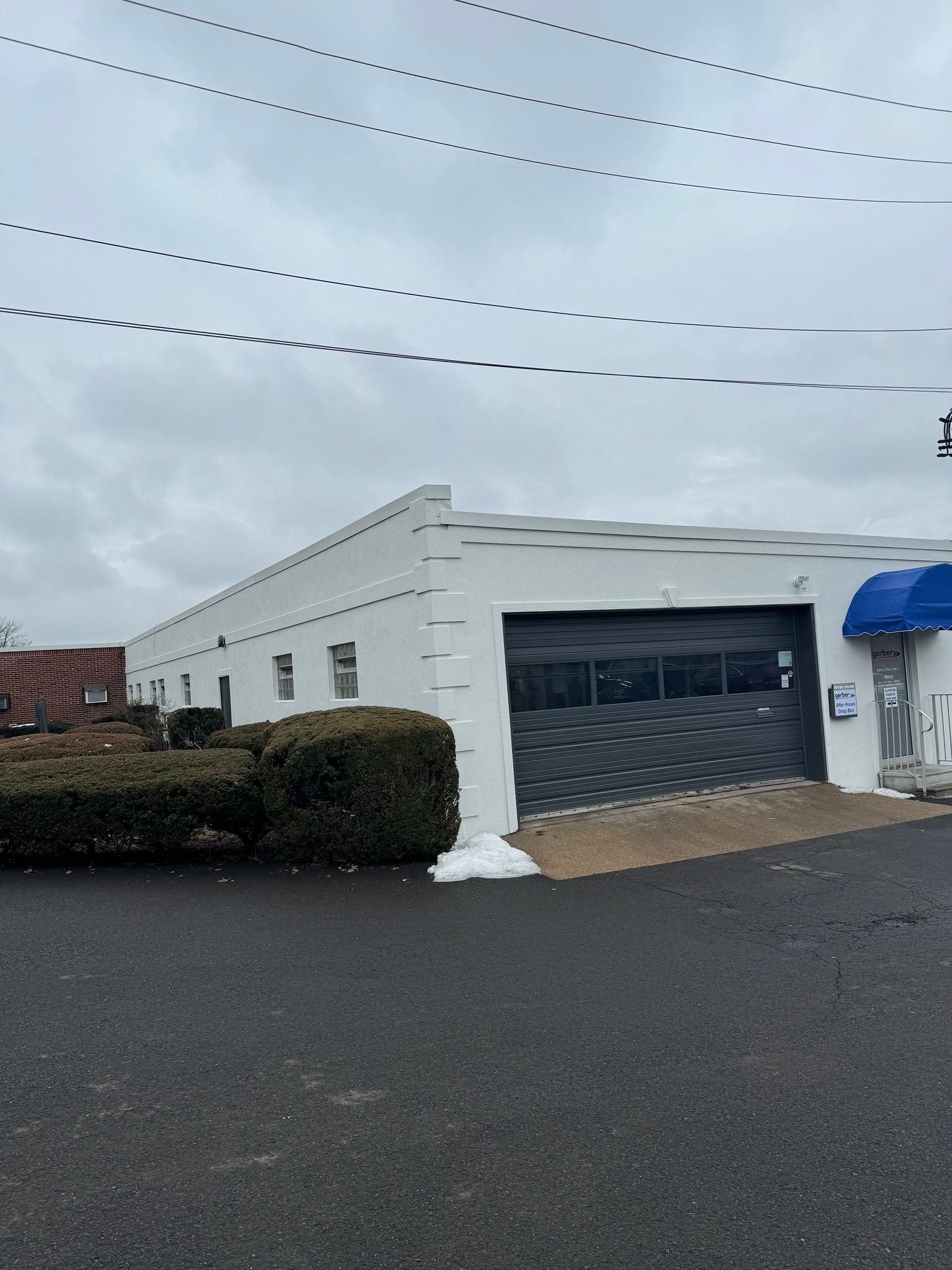 White building with a gray garage door, blue awning, and hedges under a cloudy sky.
