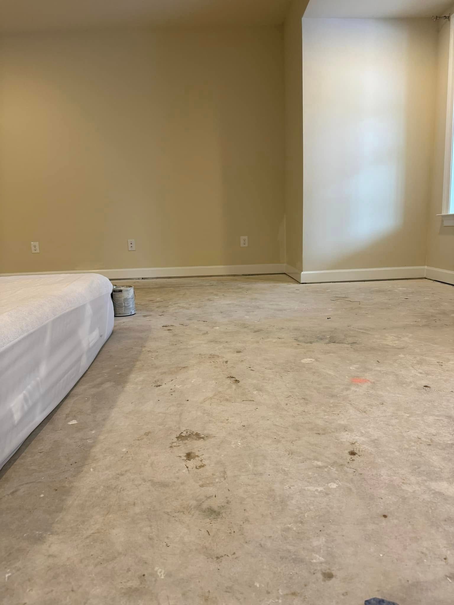 Empty beige bedroom with carpet stains and a bed partially visible on the left.