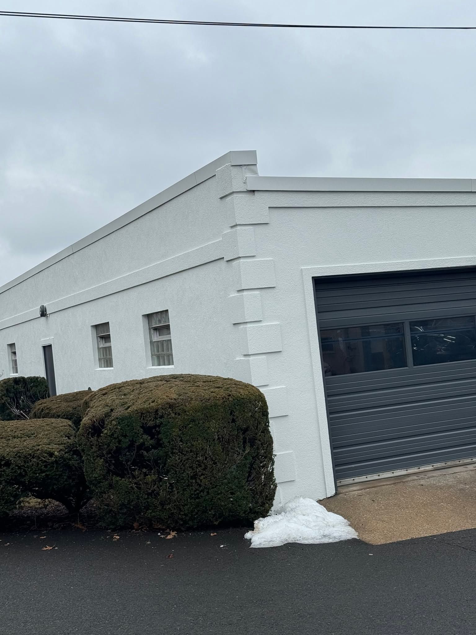 White commercial building with a black garage door and trimmed hedge on a cloudy day
