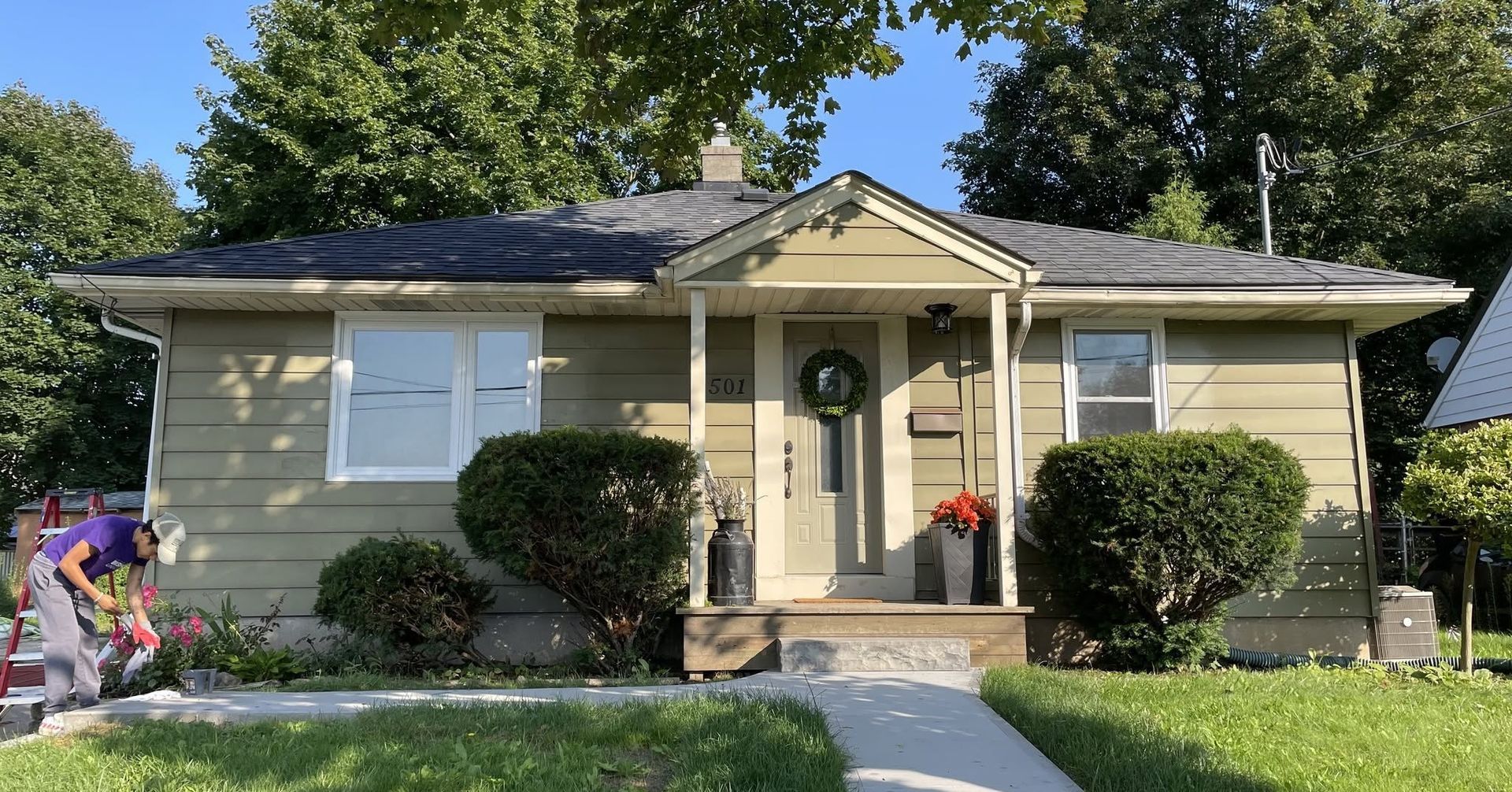 Small beige house with a front porch, green lawn, and flowerbeds in a sunny suburban yard.