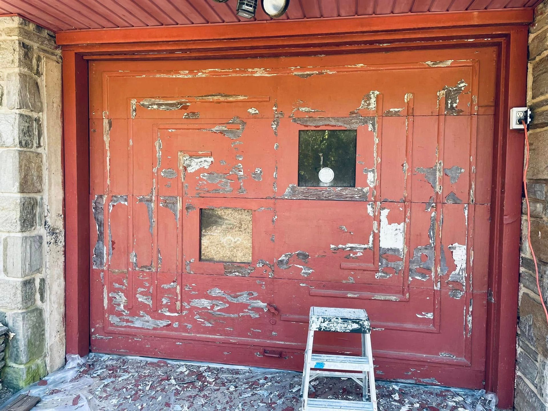 Worn red garage door with peeling paint and a small step stool in front under a wooden overhang