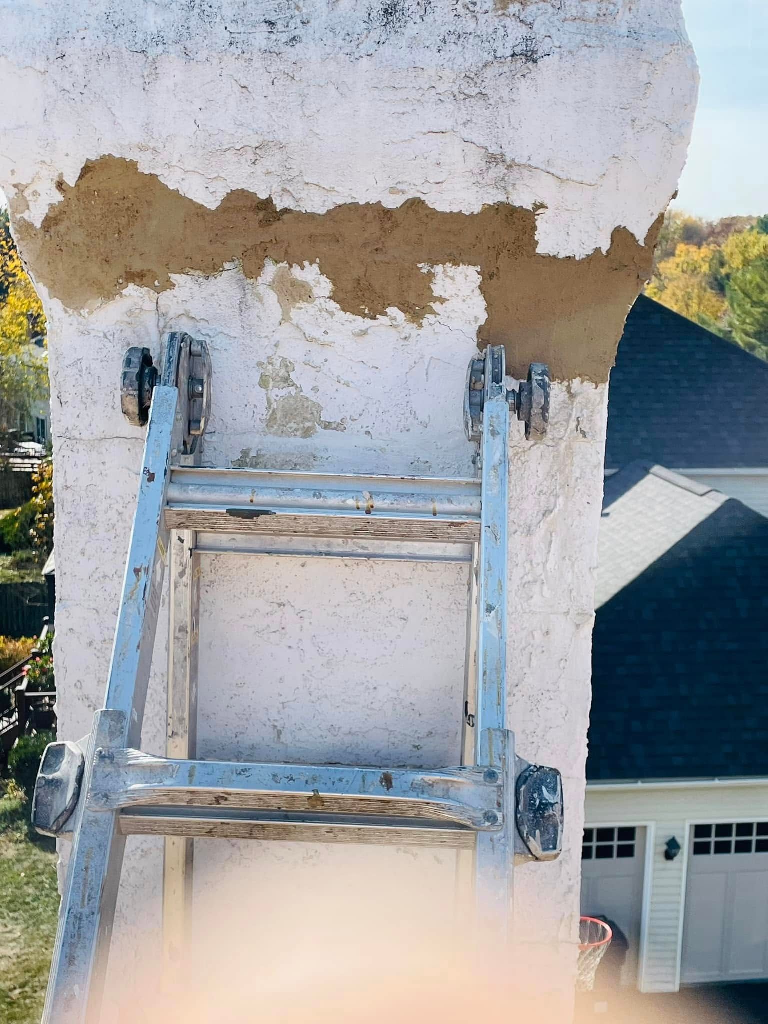 Gray ladder leaning against a weathered white wall near a house roofline, with a blurry light foreground object.