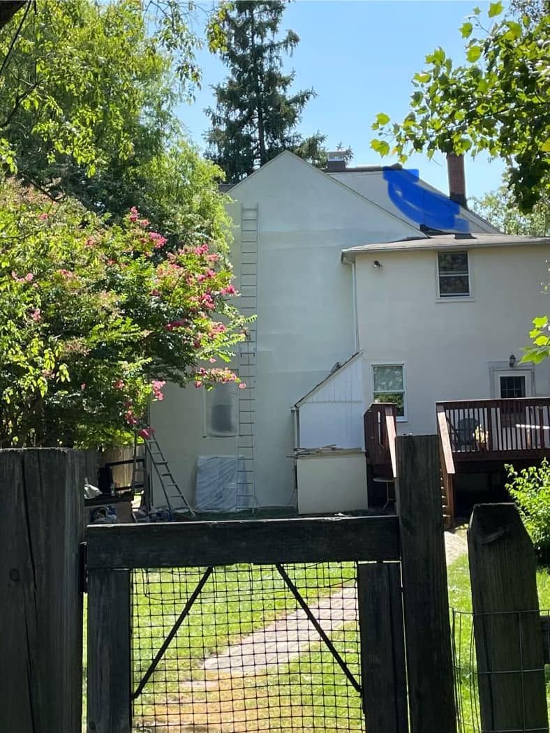 White house with blue tarp on roof behind a black gate and leafy trees