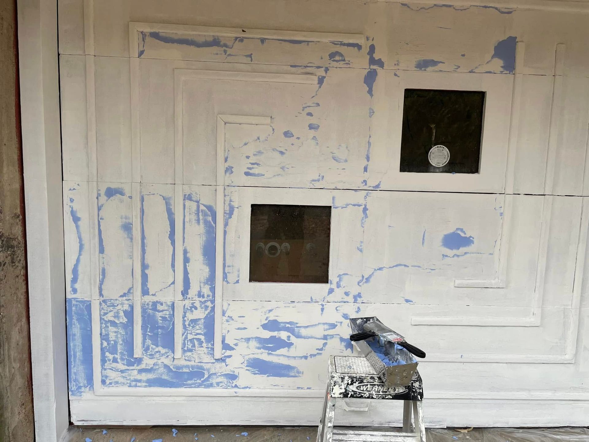 White wall with blue paint marks and two square openings, with a small paint can on the floor.