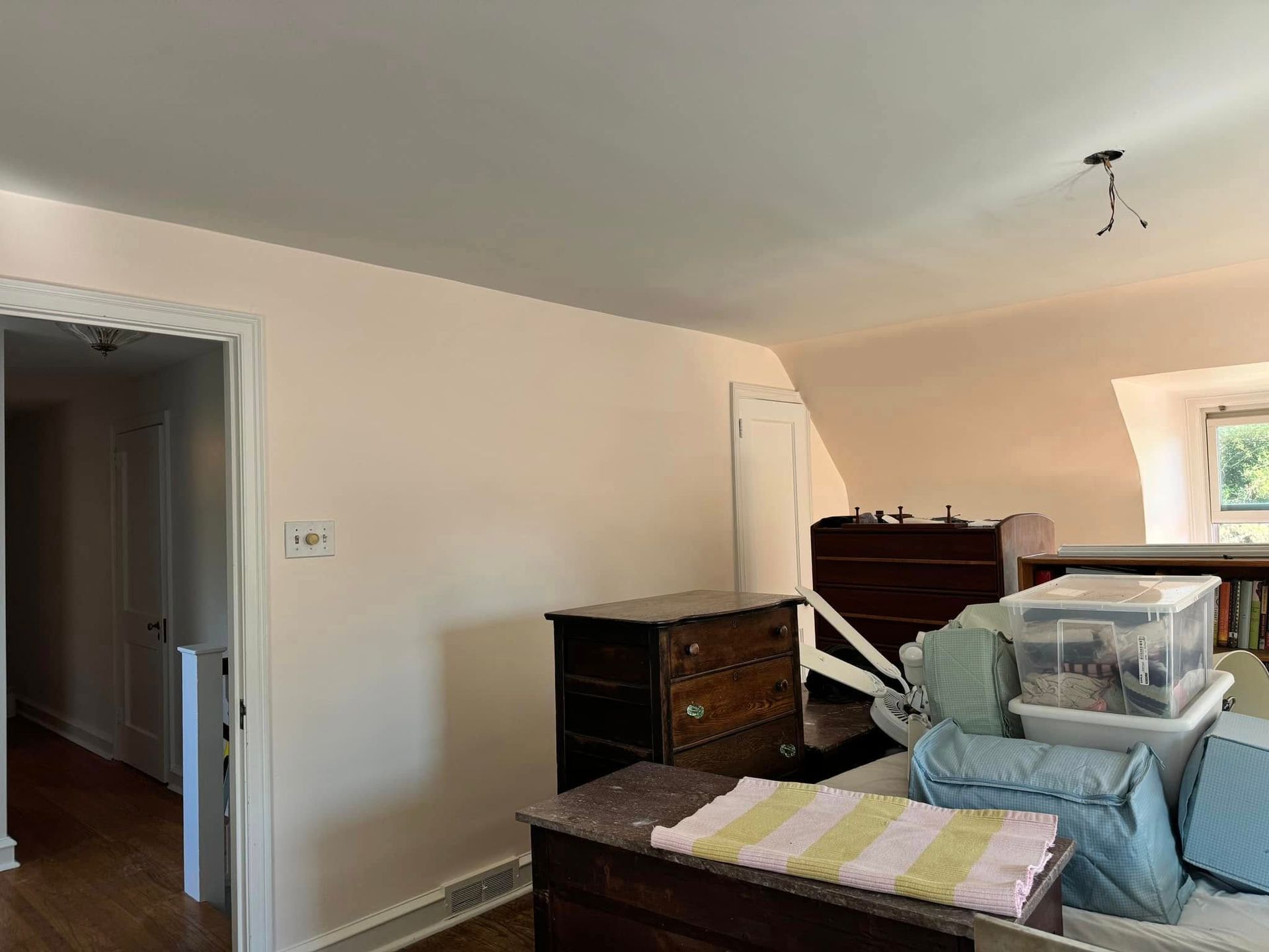 Cluttered bedroom with furniture, boxes, and a bed in a beige room near an open doorway.