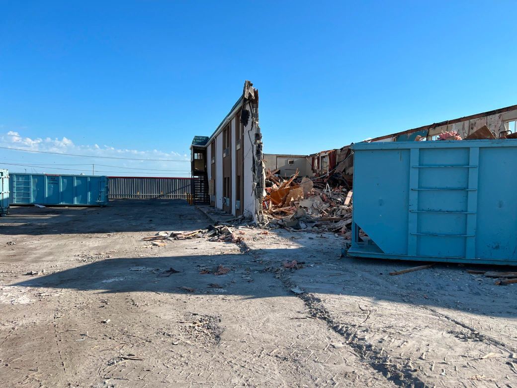 A large blue dumpster is sitting in the middle of a dirt field next to a building being demolished.