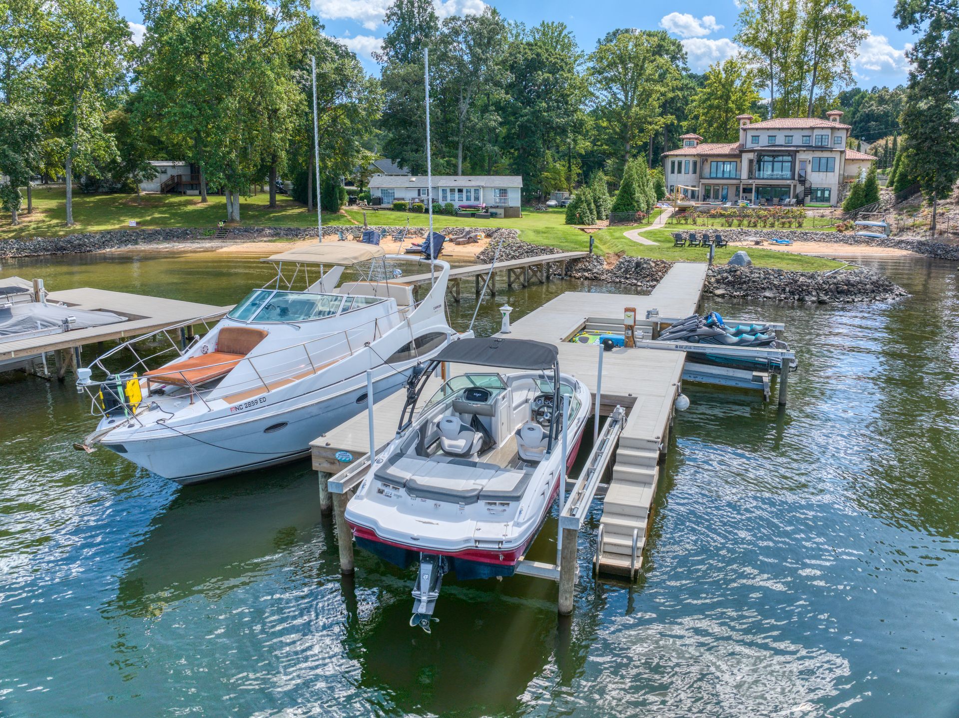 Boats docked at a pier on a lake. Luxurious home and trees in the background. Blue water and sky.