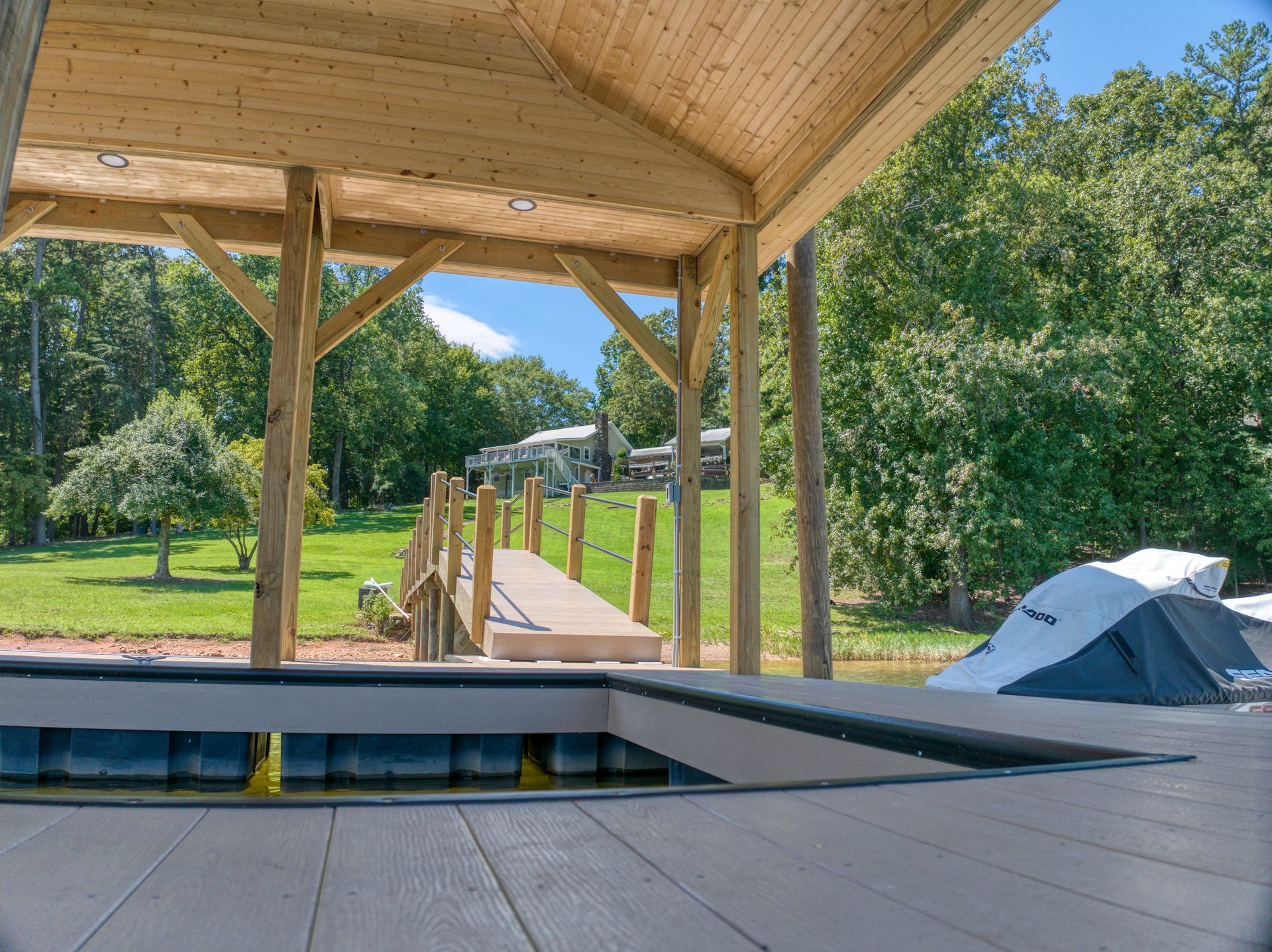 Dock with wooden beams, roof, leading to a house in the distance, blue sky, and green trees.