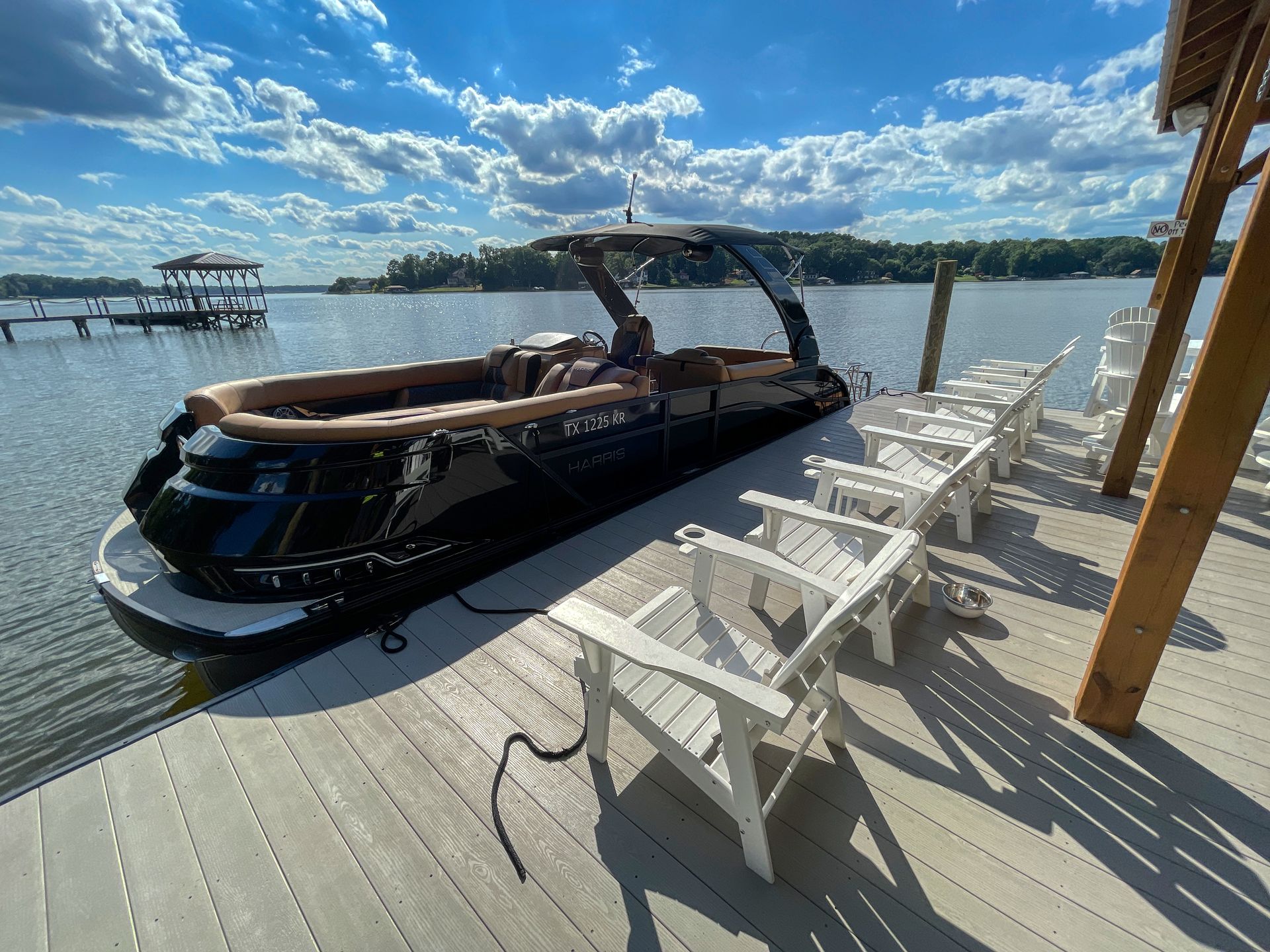 Black pontoon boat docked on a wooden pier with white chairs, lake view, sunny day.
