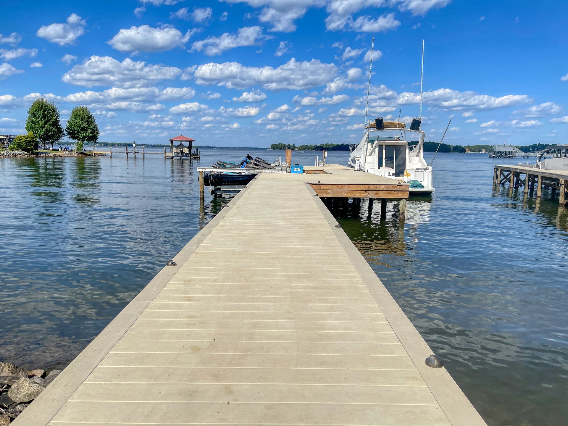 A long concrete dock leads to a white boat on calm water; blue sky with puffy clouds in background.