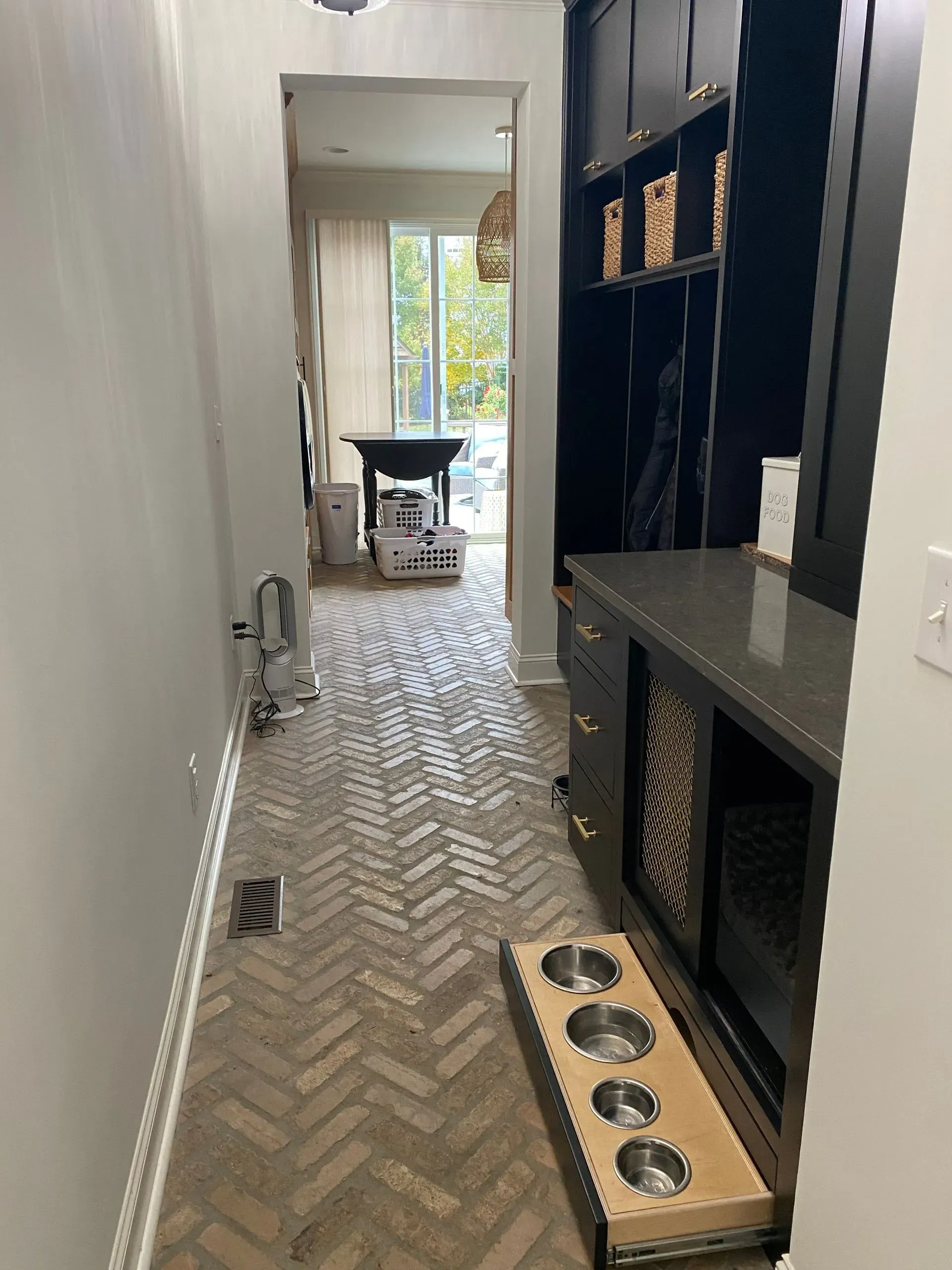 Hallway with herringbone brick floor, black built-in storage, dog bowls, and a view to a dining area.