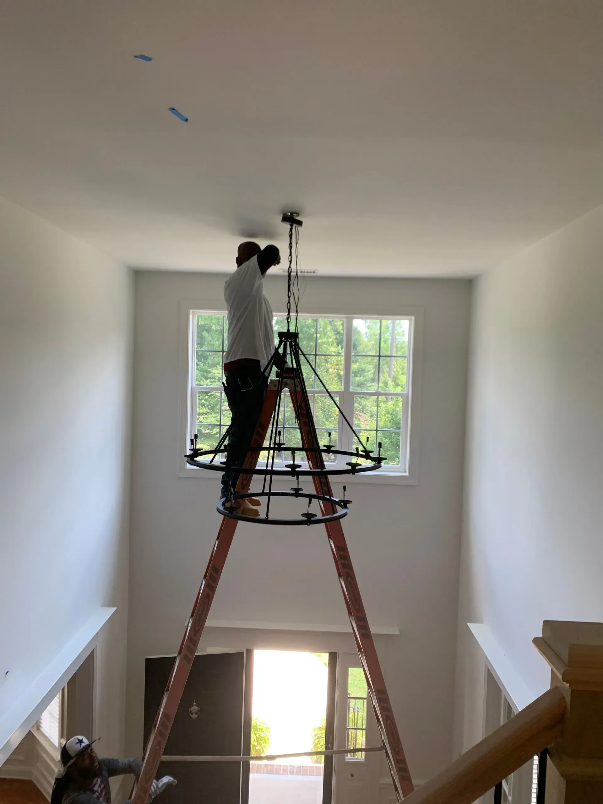 Man installing a chandelier on a tall ladder in a white-walled room with a large window and a doorway.