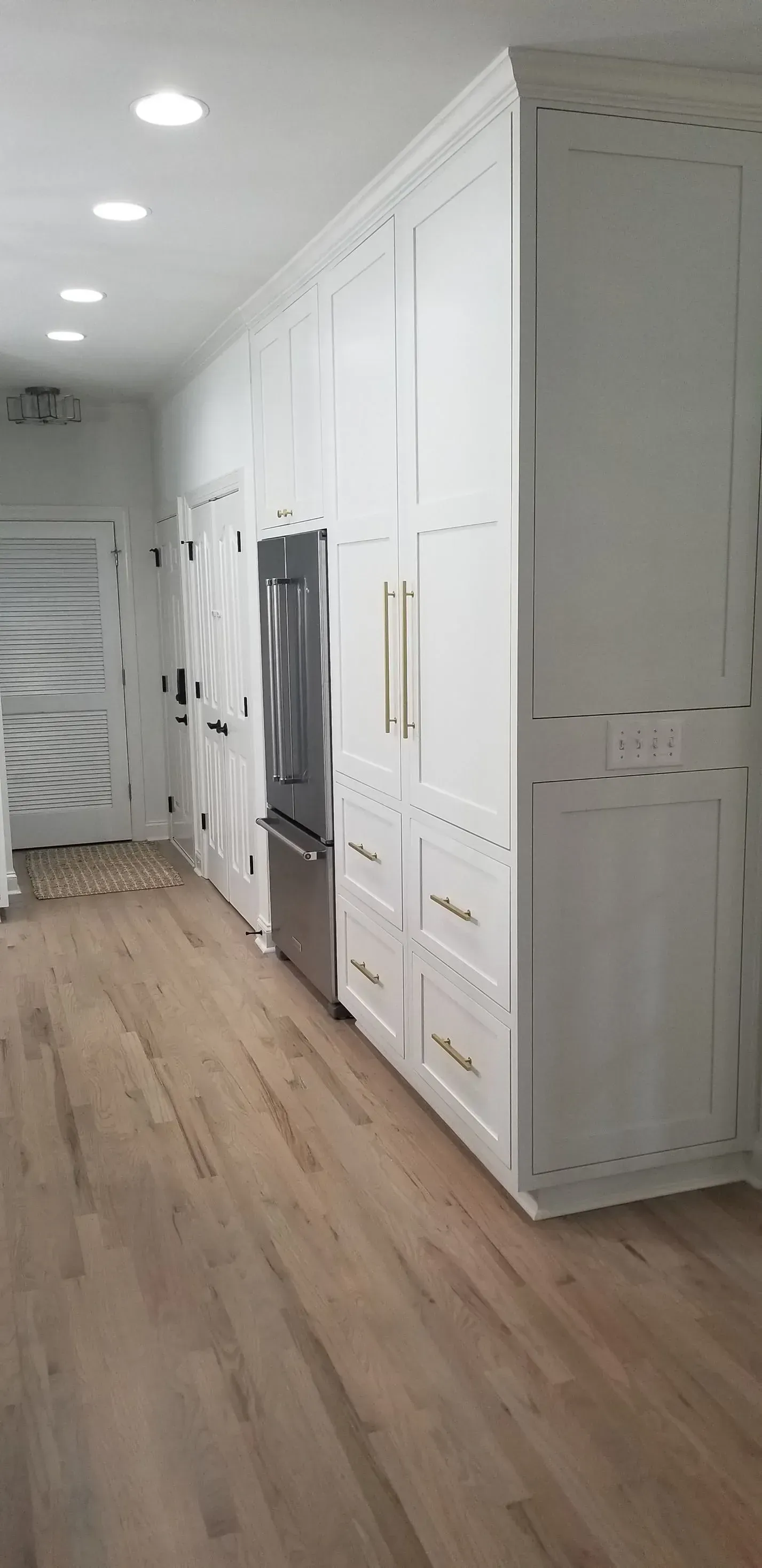 A white kitchen with tall cabinets, a stainless steel refrigerator, and hardwood flooring.