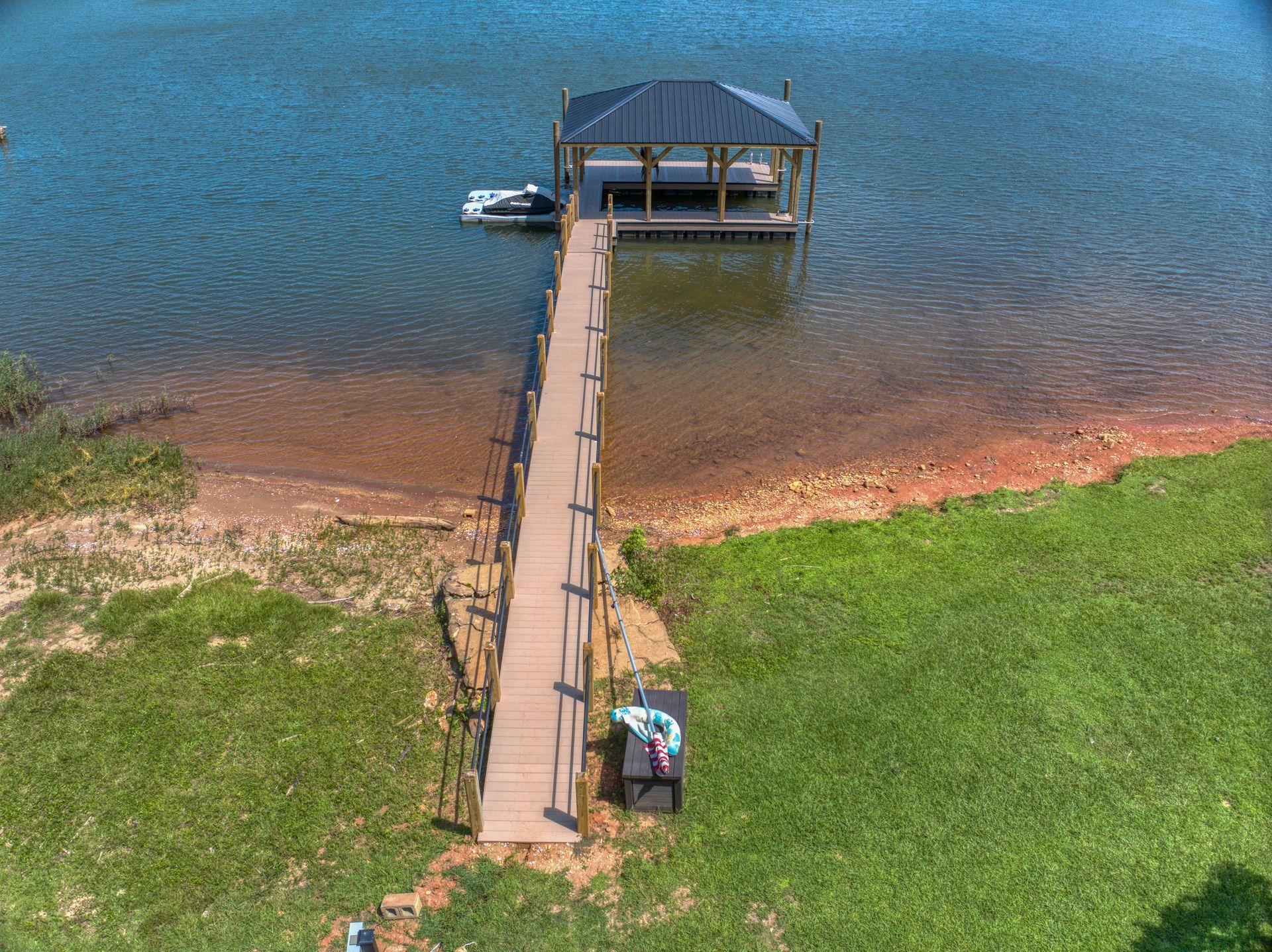 Dock with a covered boat slip extends into a blue lake; grassy shoreline on right, brown dirt and reddish shore on the left.