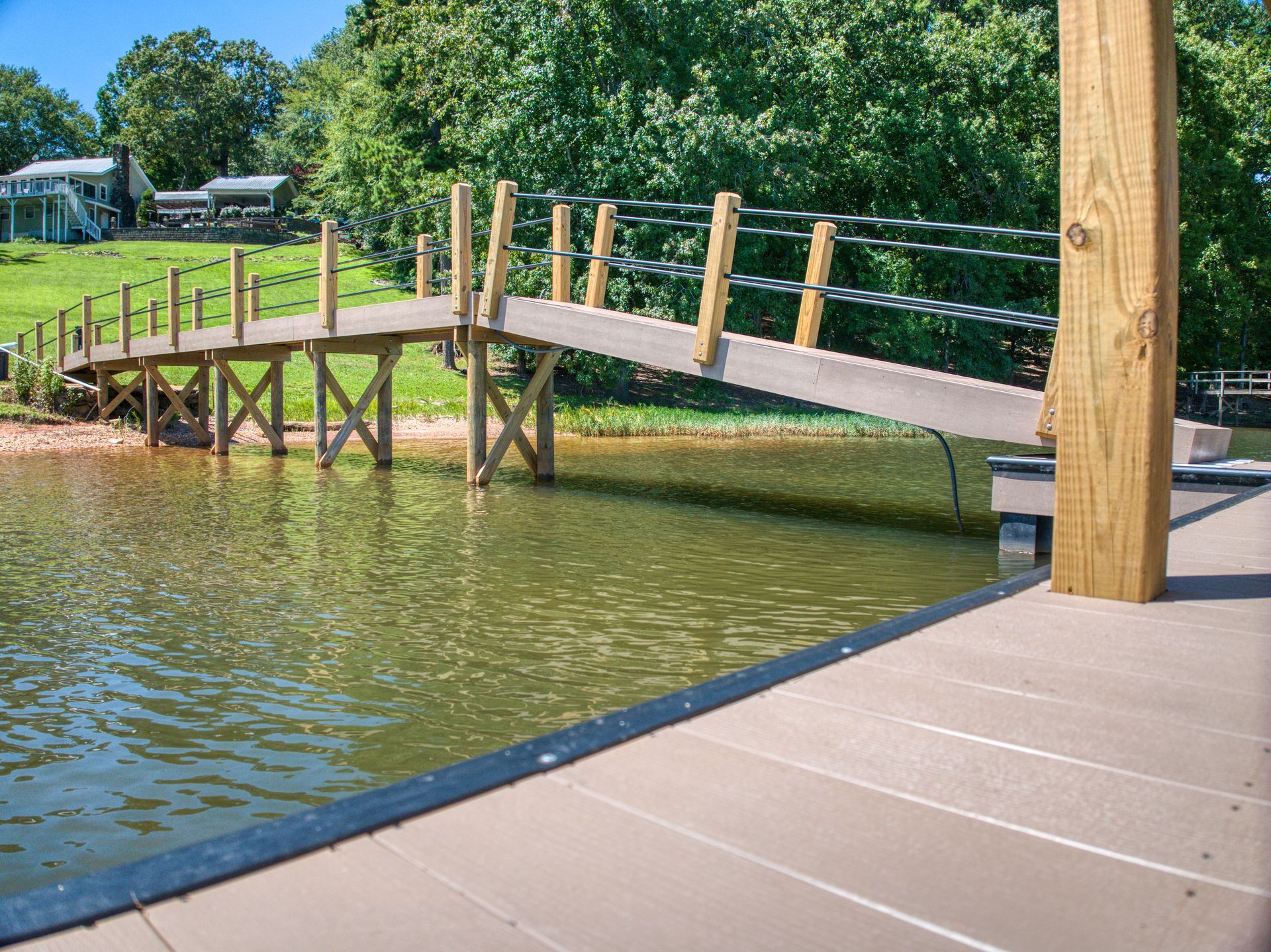 Wooden bridge over water, connecting a dock to a grassy bank with a house in the background.