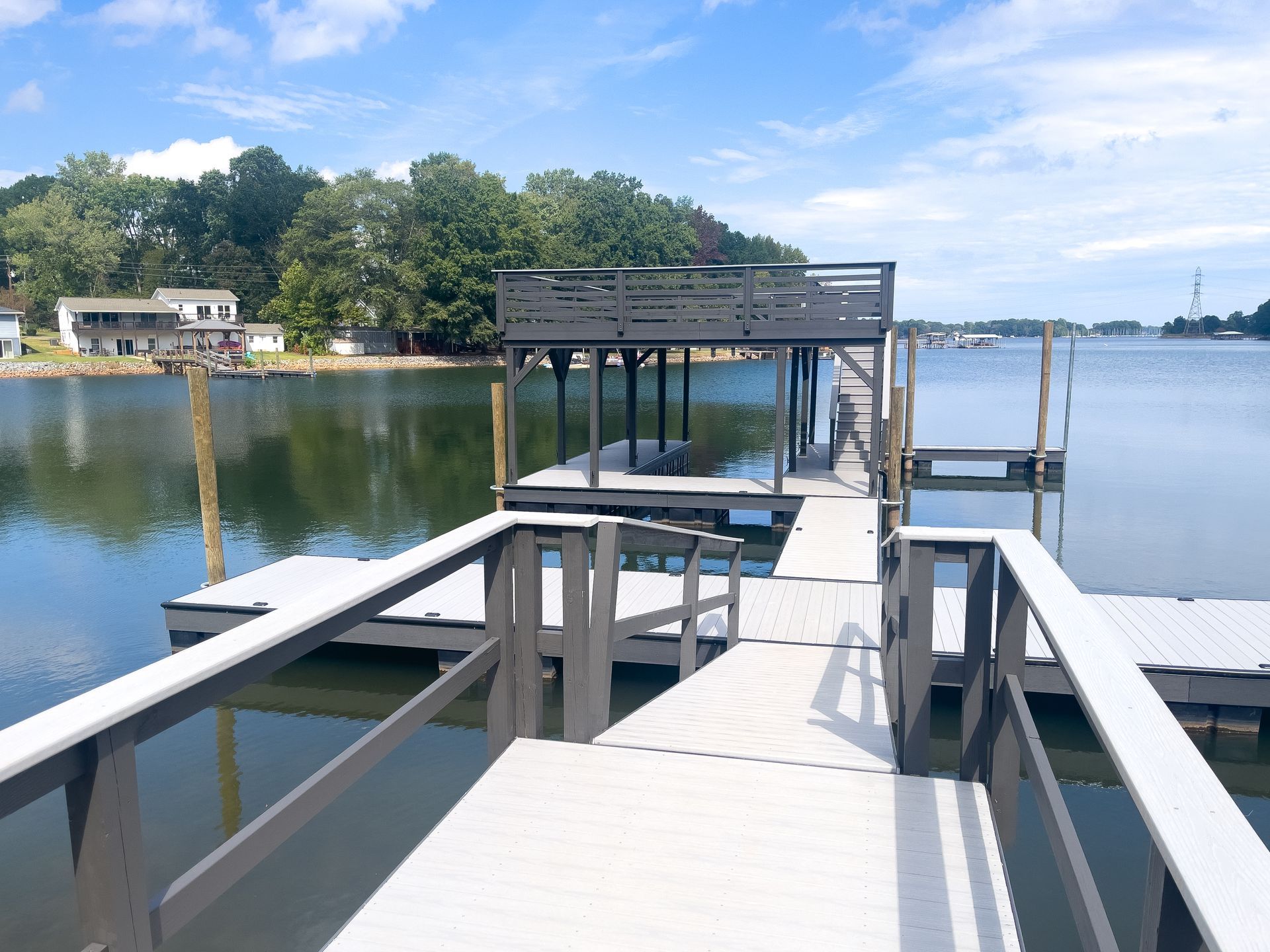 Dock on a lake with a dark gray upper platform and white walkways. Houses and trees are visible in the background.