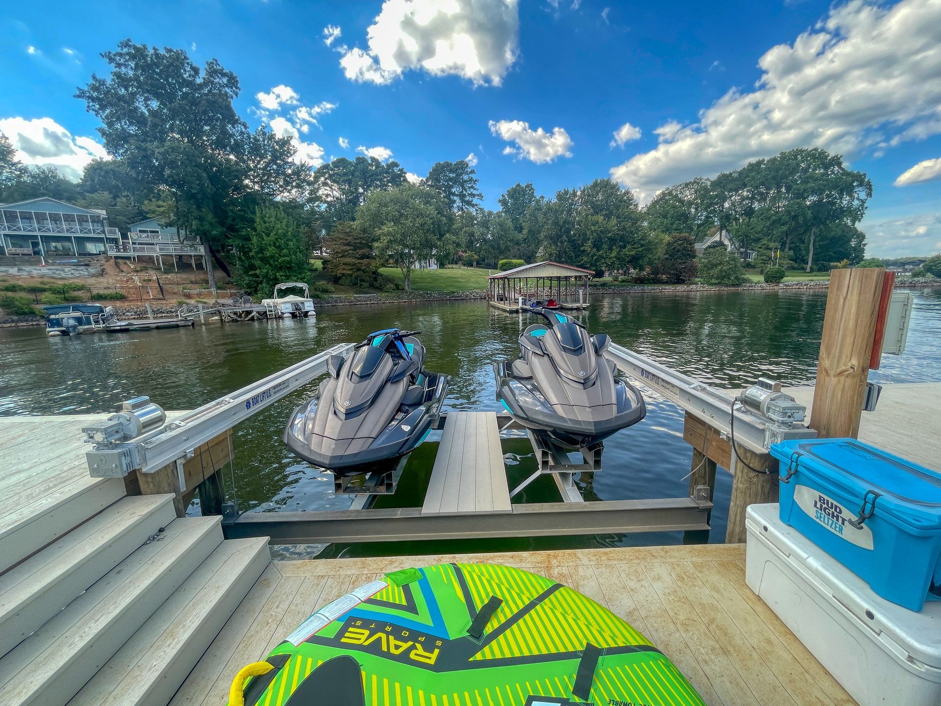Two jet skis docked on a lift, waterfront view. Blue skies, trees, wakeboard, and lake.