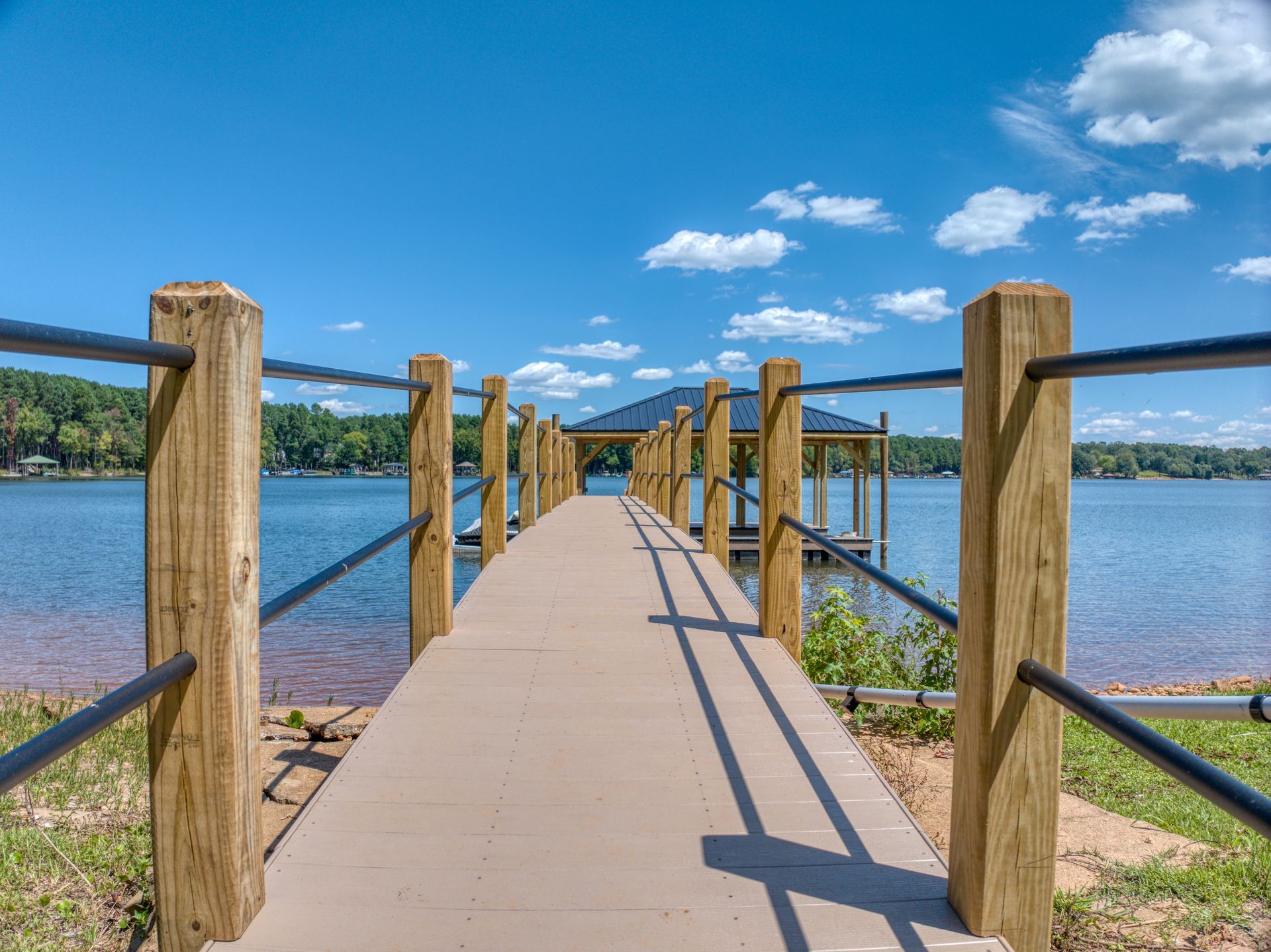 Wooden pier extending into a calm lake, leading to a covered dock under a bright blue sky with fluffy clouds.