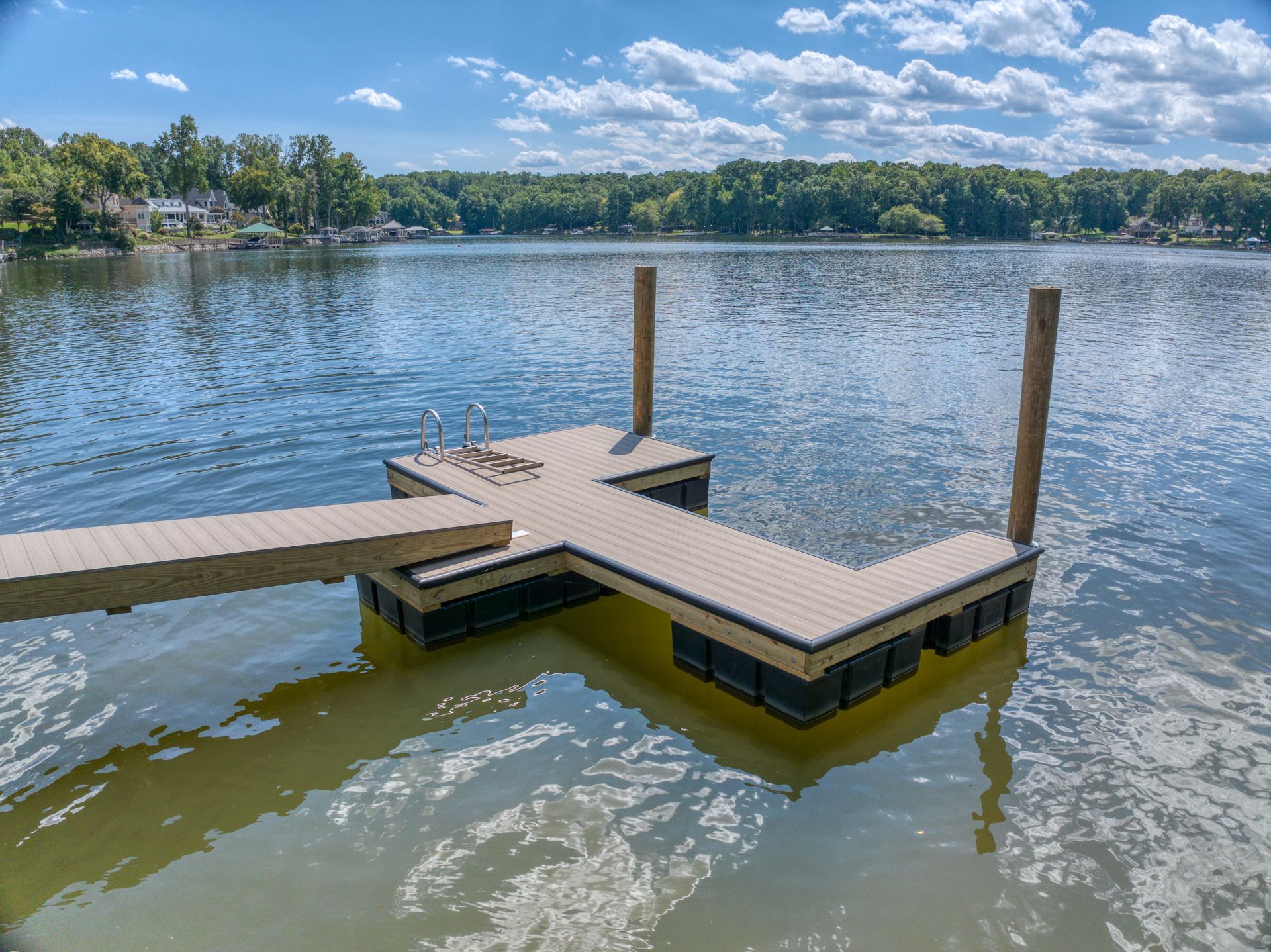 Floating dock on a lake with a wooden walkway and posts. Trees and houses in the background. Sunny day.