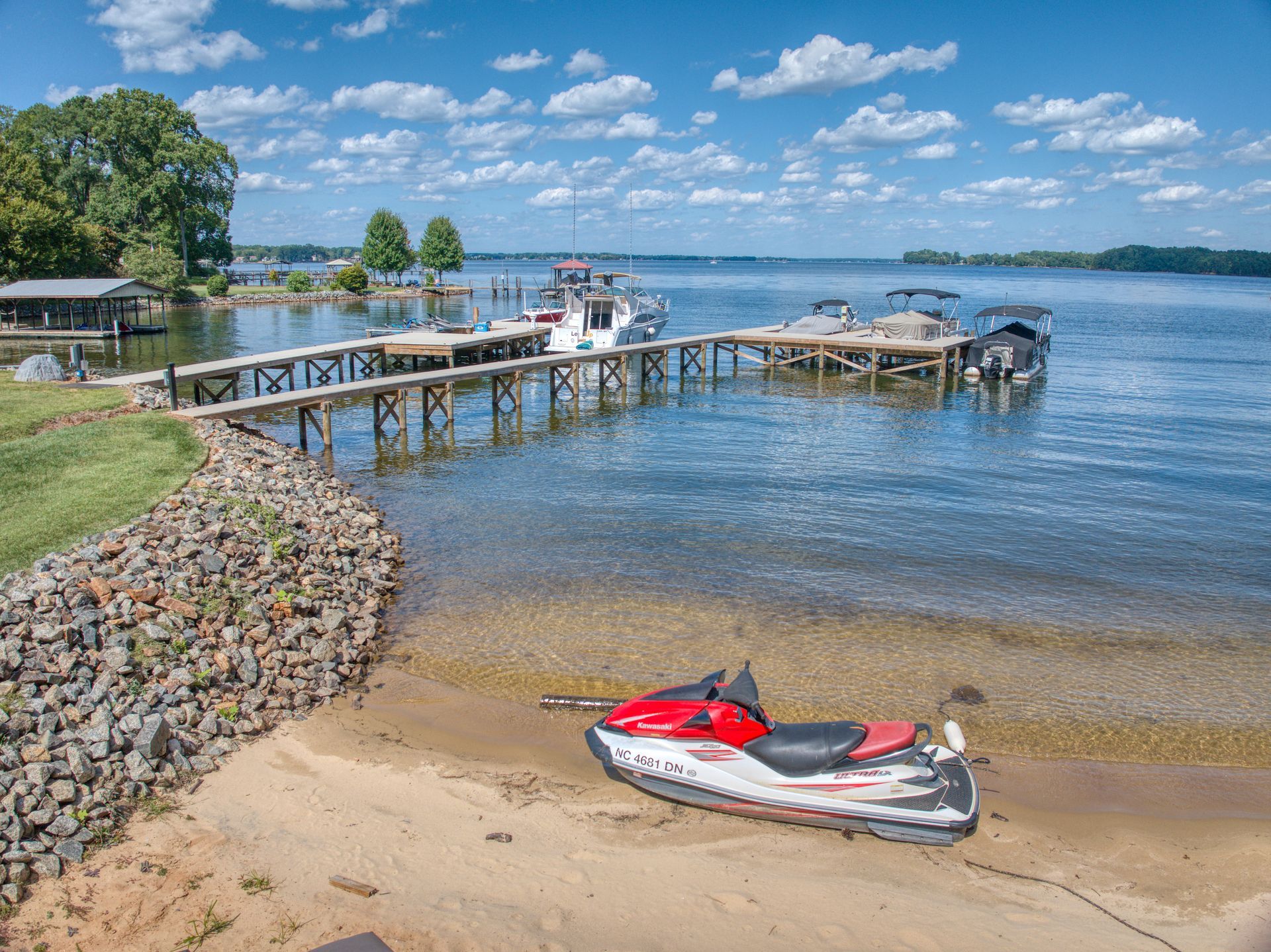 Jet ski on a sandy beach in front of a lake with docks and boats under a blue sky with clouds.